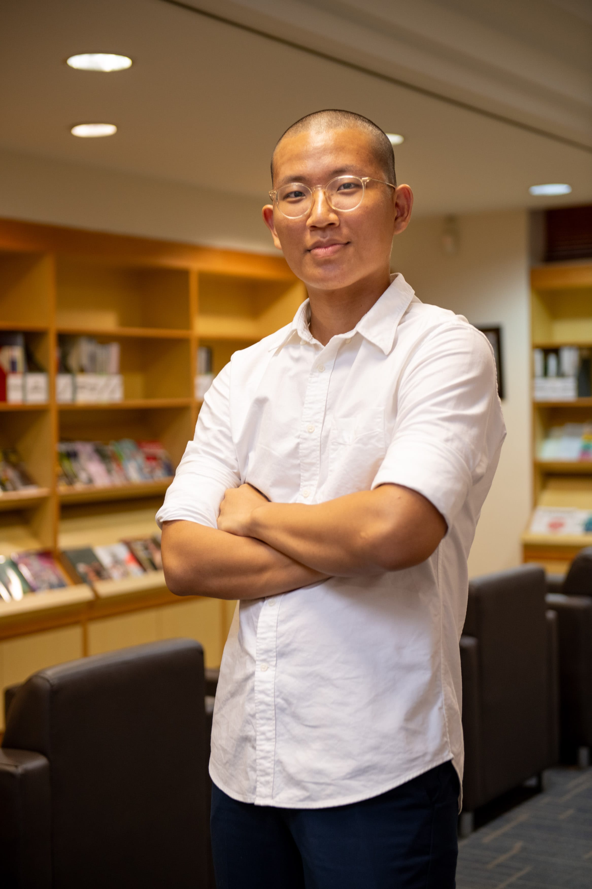 A man with a shaved head and clear-framed glasses stands arms crossed in a library, wearing a white shirt.