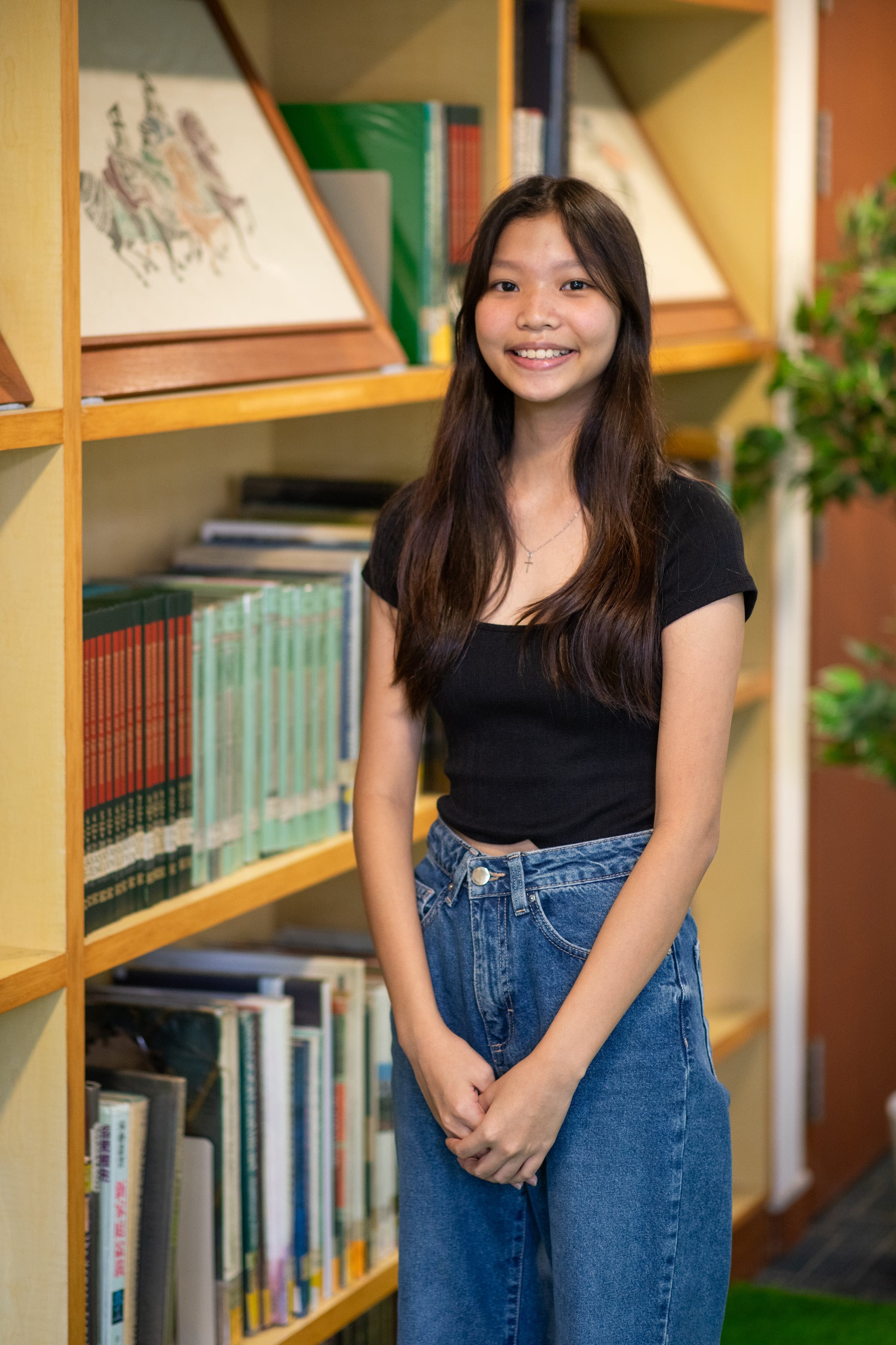 Young woman with long dark hair, black top, and blue jeans smiling in front of wooden bookshelves filled with books.