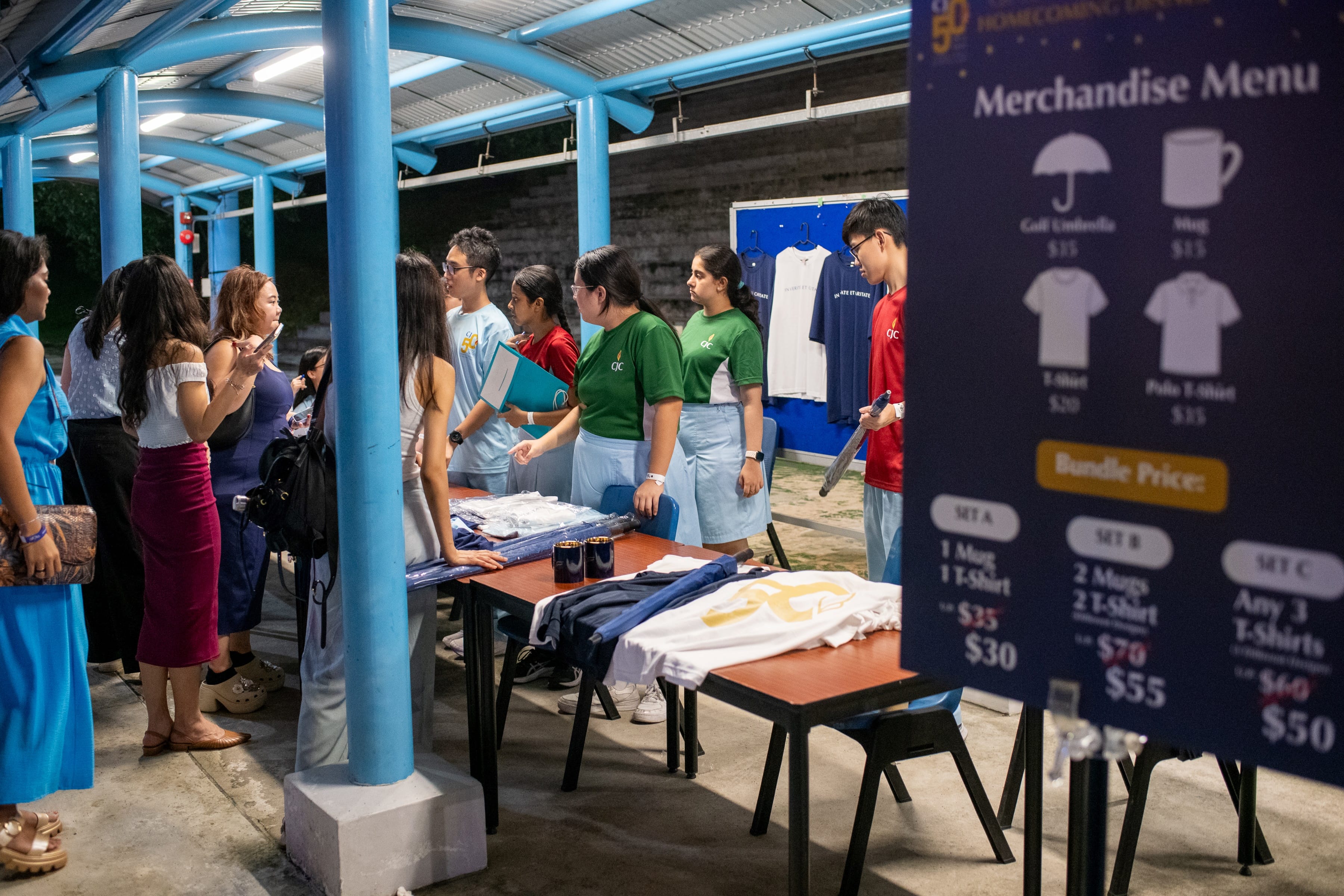 People browse a merchandise booth at a homecoming event, with a navy menu board listing t-shirts, mugs, and bundle prices.