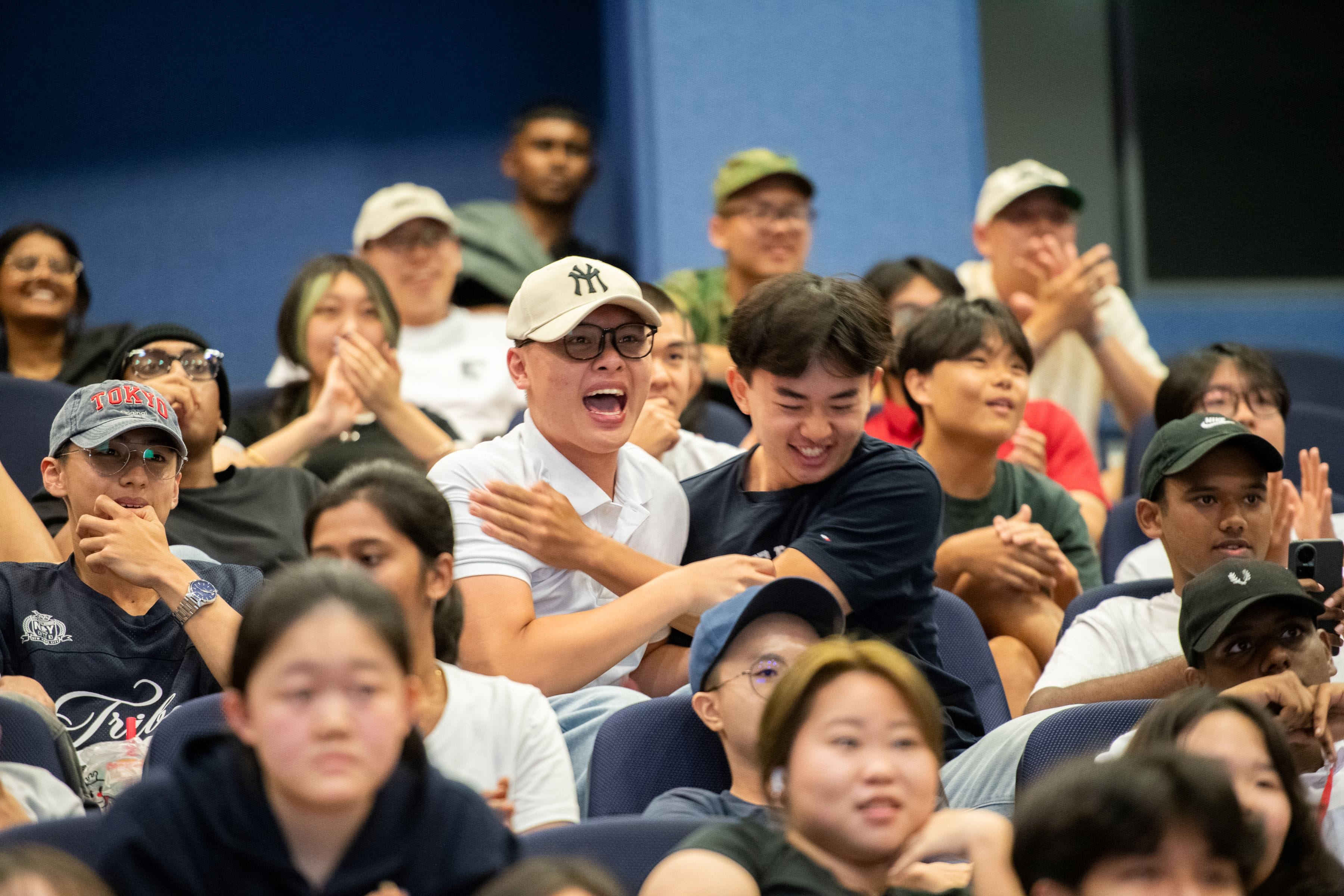 A large crowd of young people seated in an auditorium, many clapping and cheering with excited expressions.