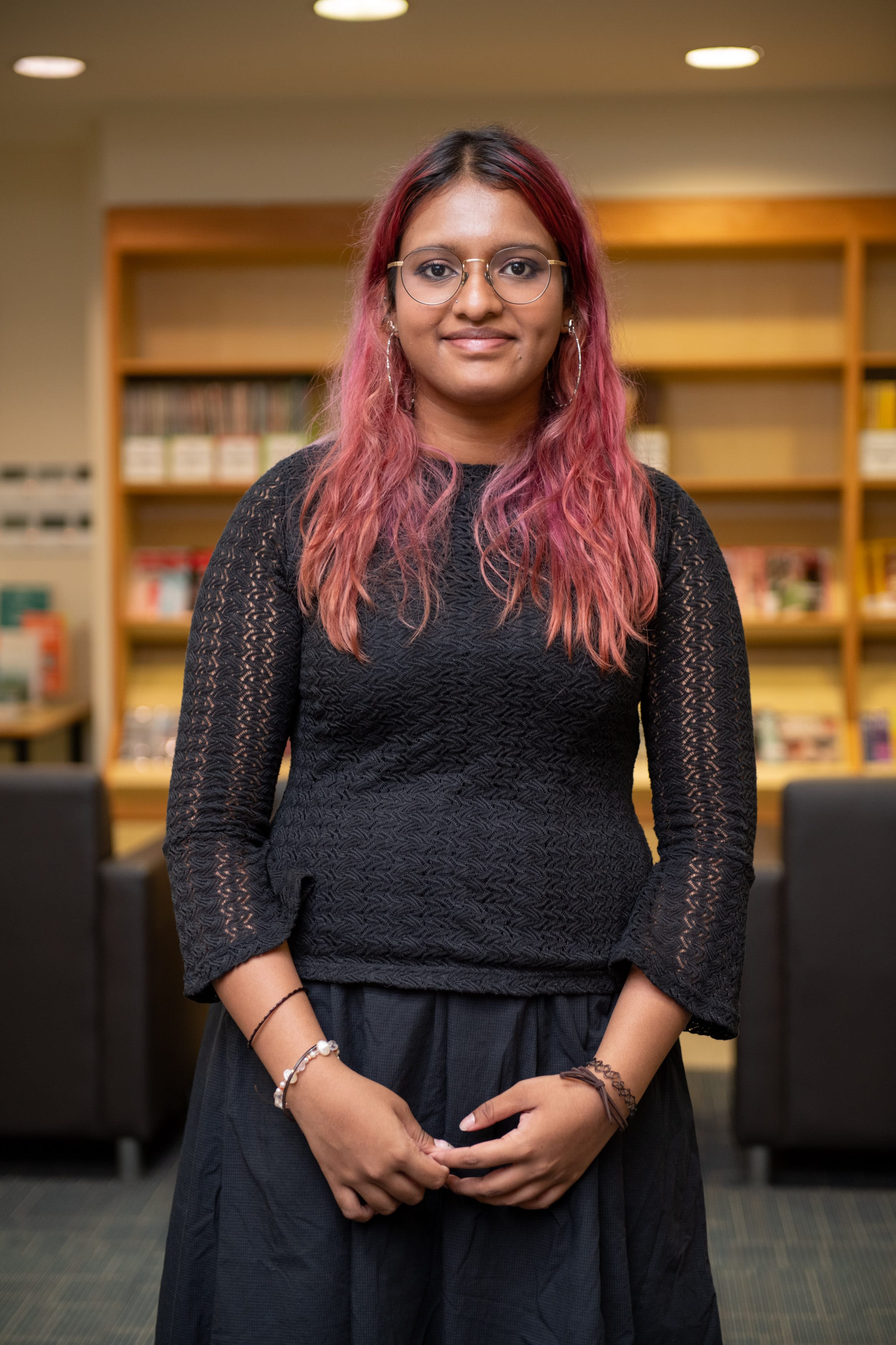Young woman with pink hair and glasses wearing a black top, standing in a library with bookshelves behind her.