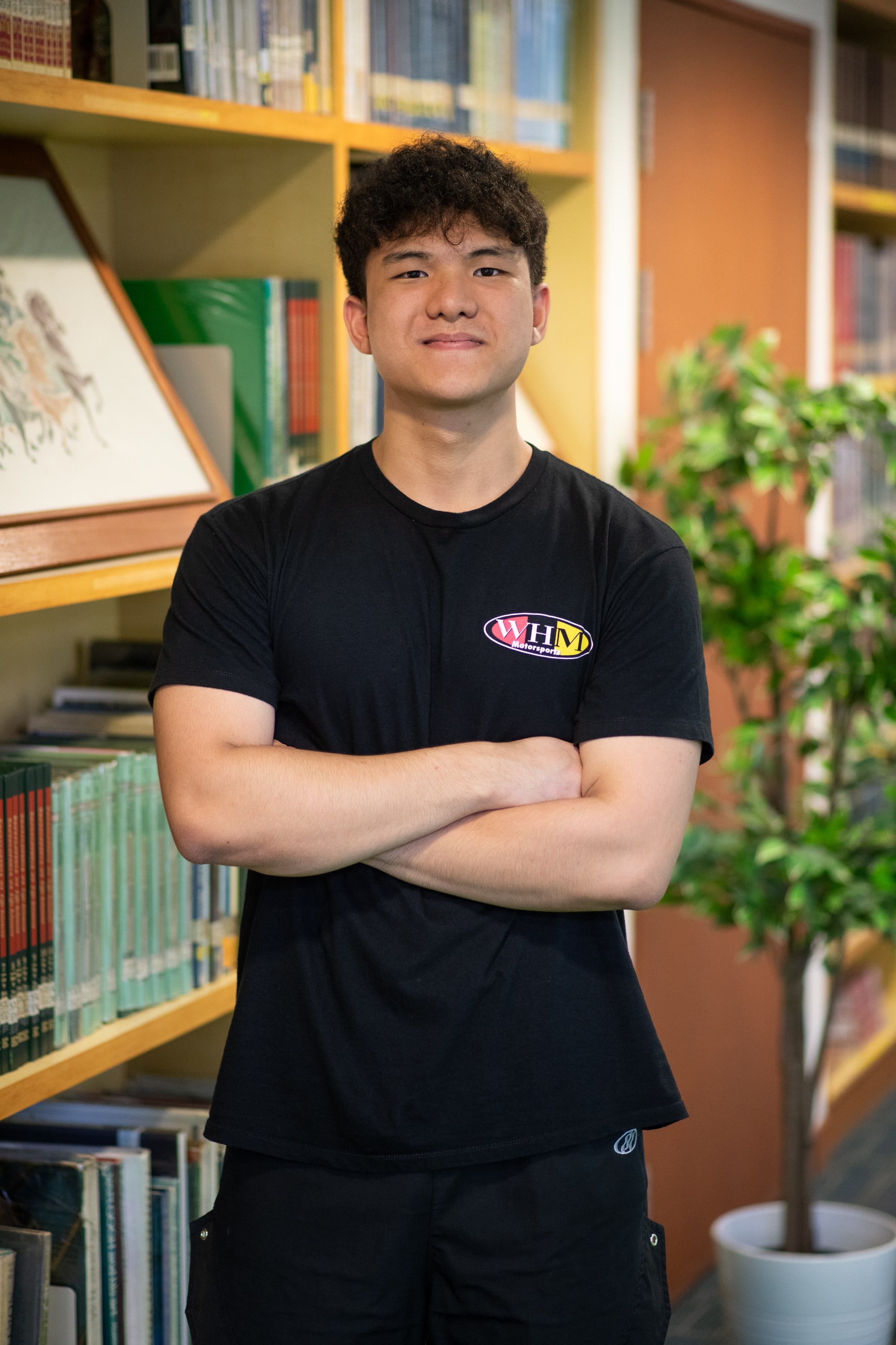 Young man with curly hair in a black WHM Motorsports shirt stands arms crossed, smiling in a library.