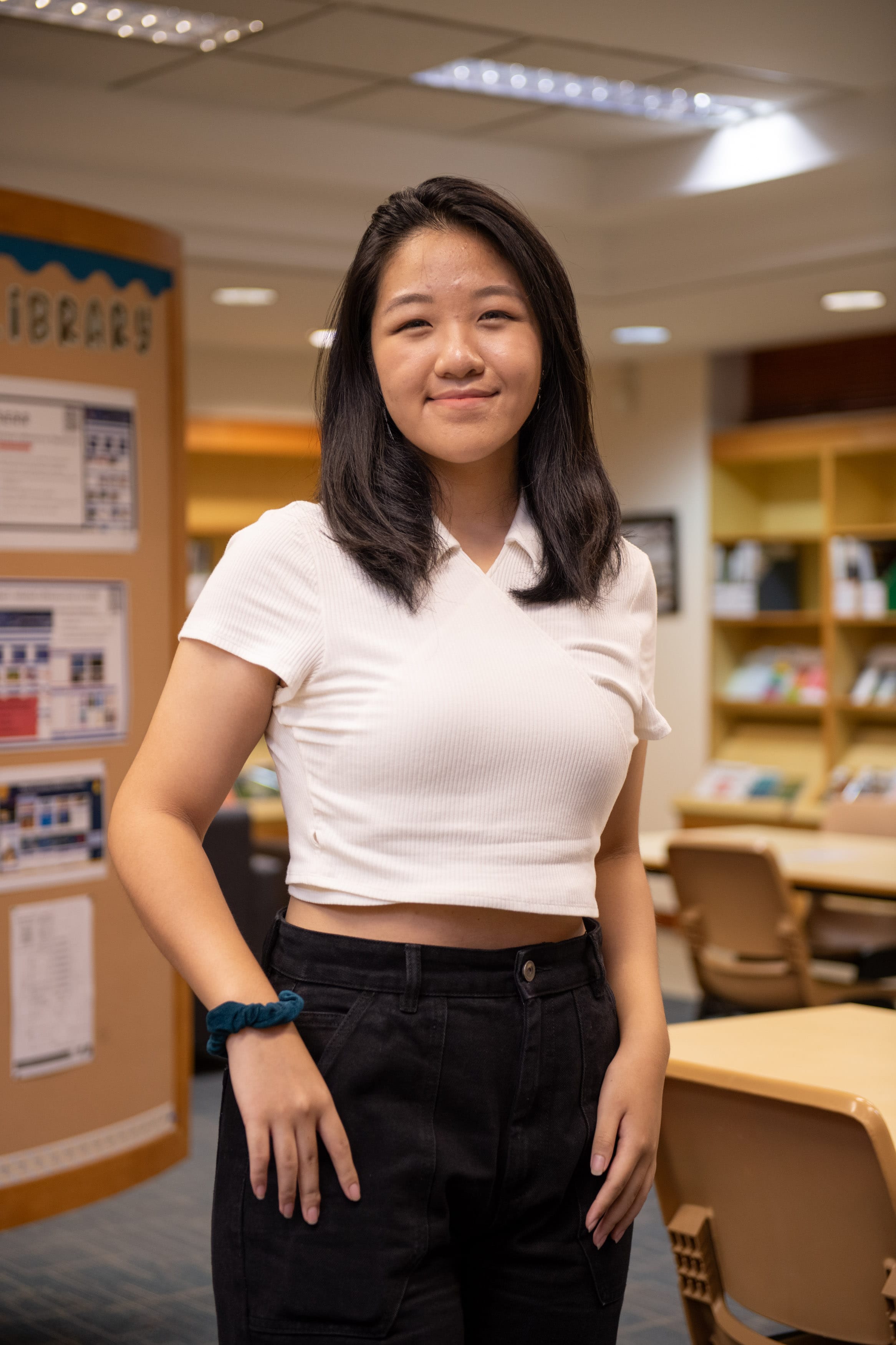 Young woman with dark hair smiling, wearing a white crop top and black jeans, standing in a school library.