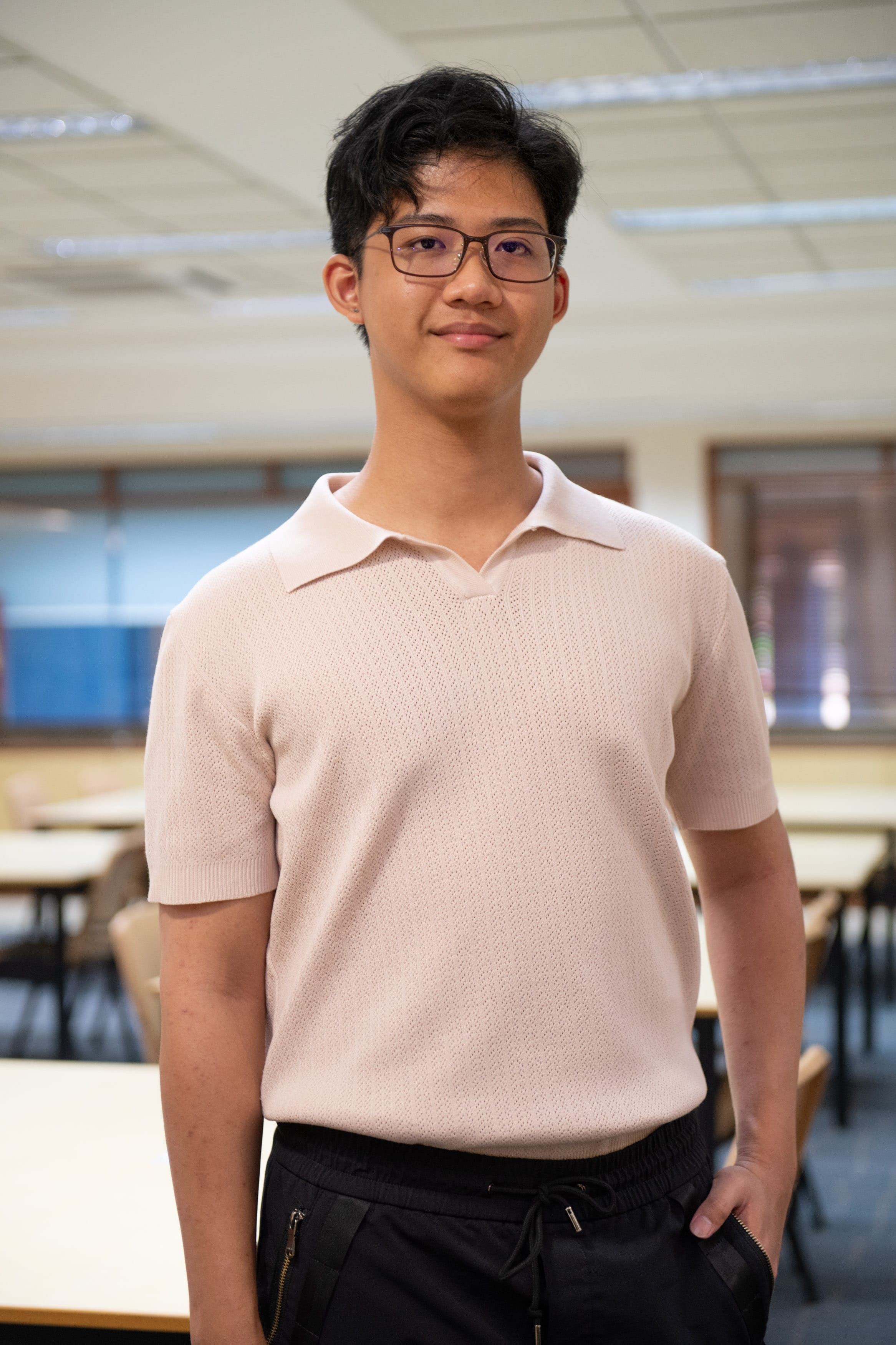 Young man with glasses and short dark hair wearing a pink polo shirt and black pants, standing in a classroom.