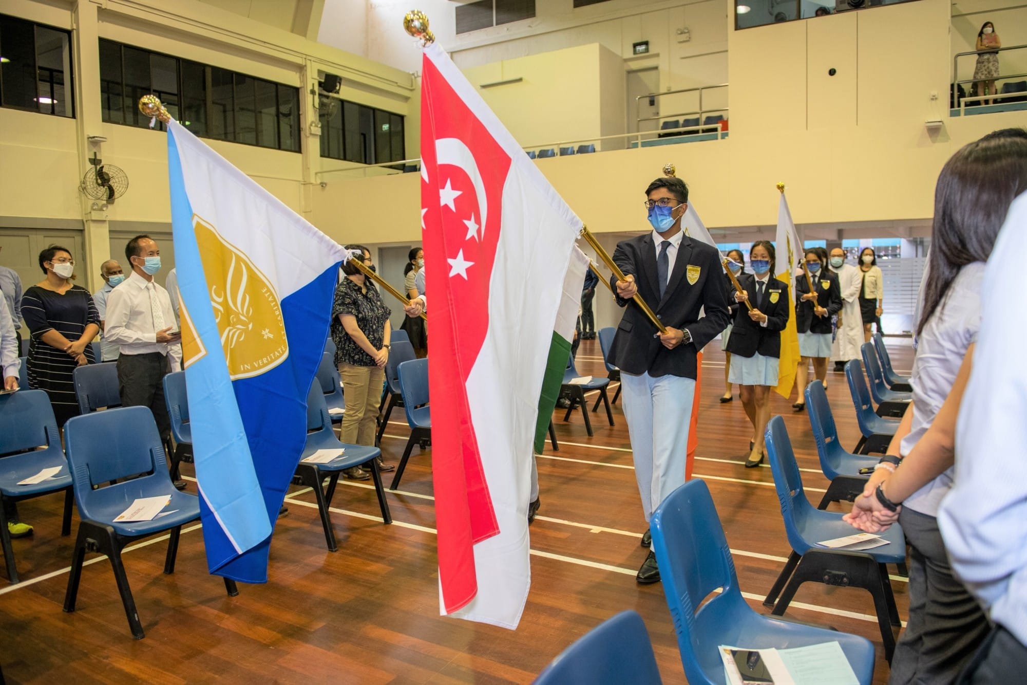 Students in blazers carry the Singapore flag and a school flag during a indoor ceremony, with masked attendees standing nearby.