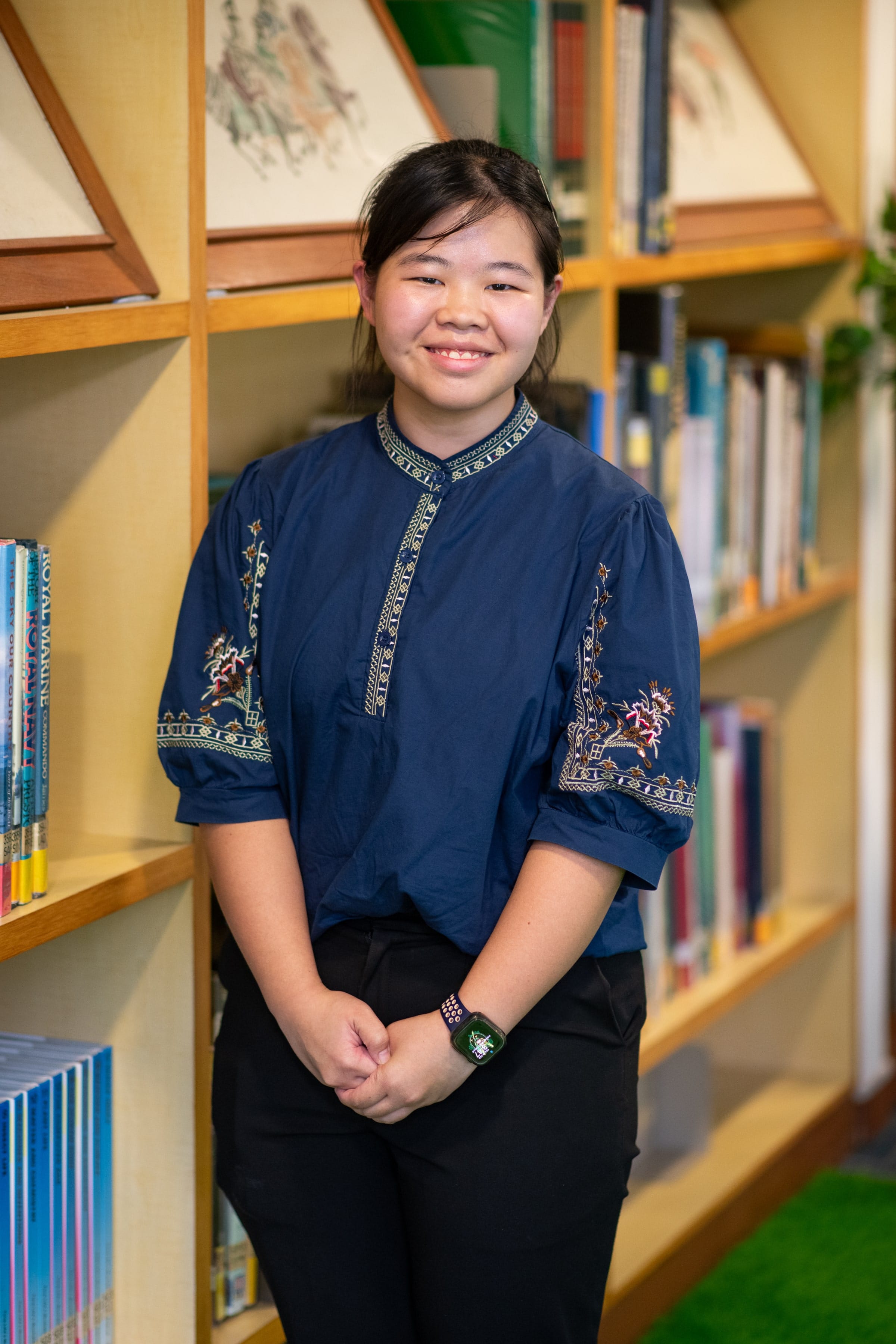 A young woman smiling in a navy embroidered top and black pants, standing in front of wooden bookshelves.