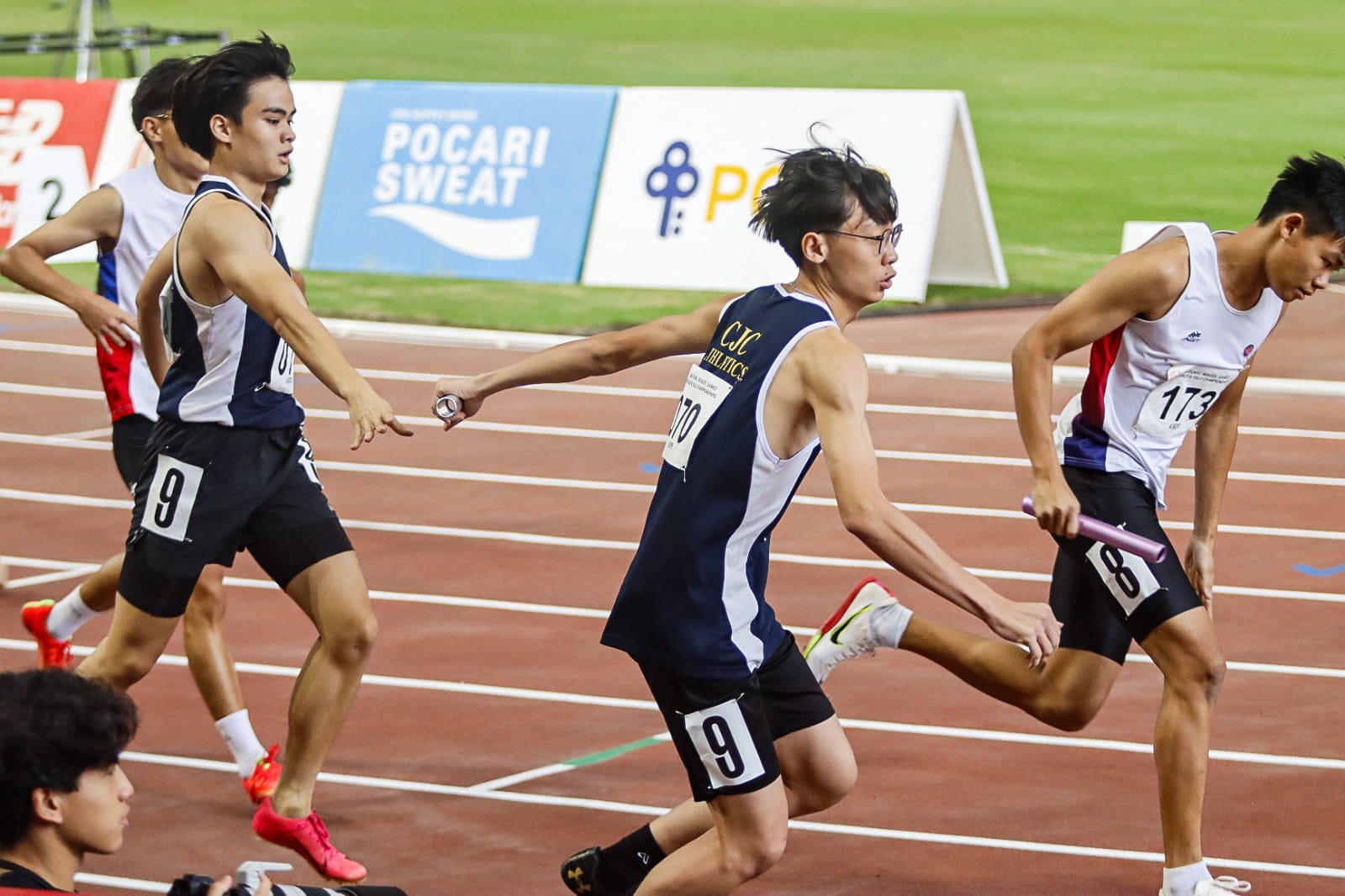 Male relay runners on a red track passing batons, with a Pocari Sweat banner visible in the background.