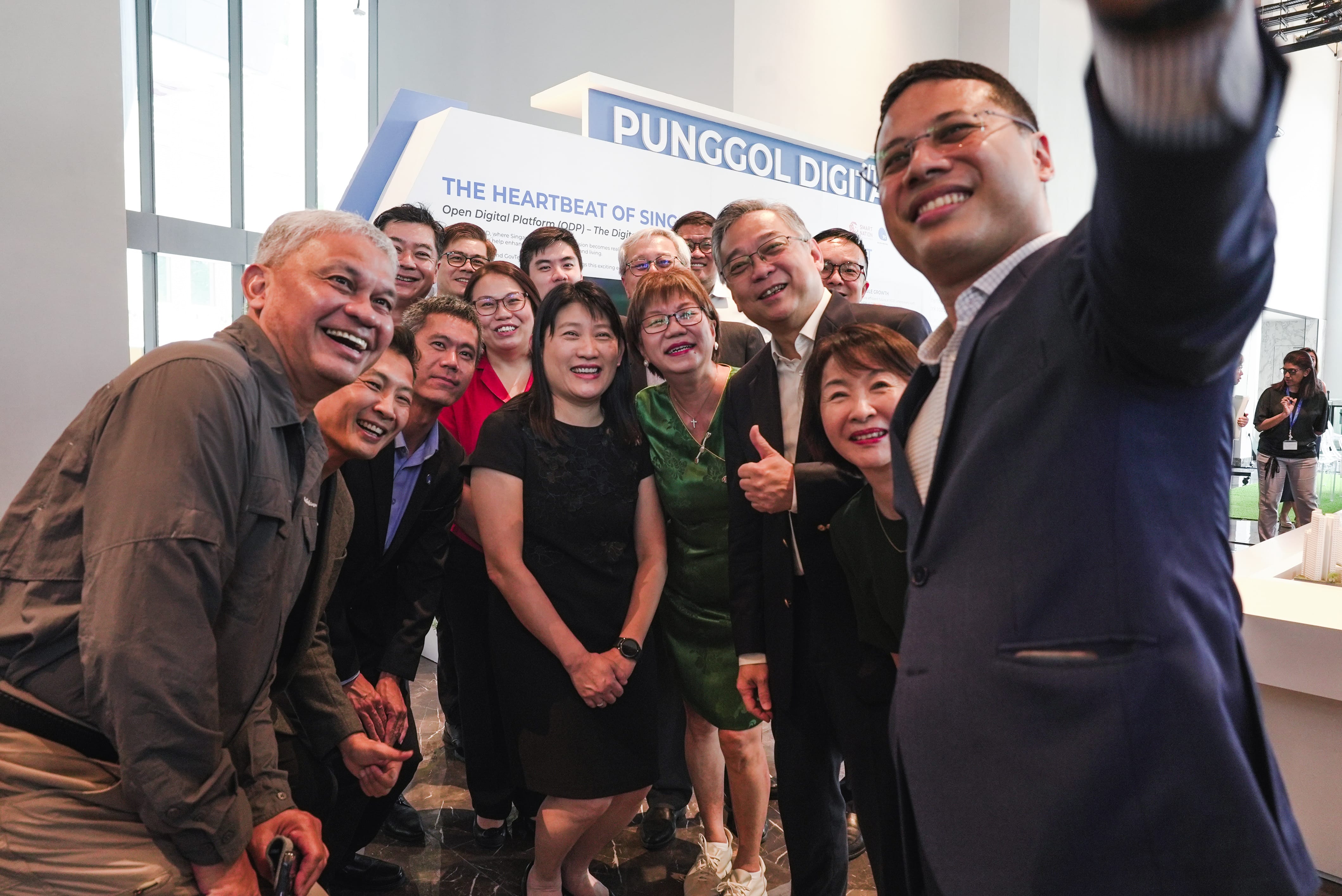 Group of smiling people pose for a selfie indoors, with a Punggol Digital sign in the background.