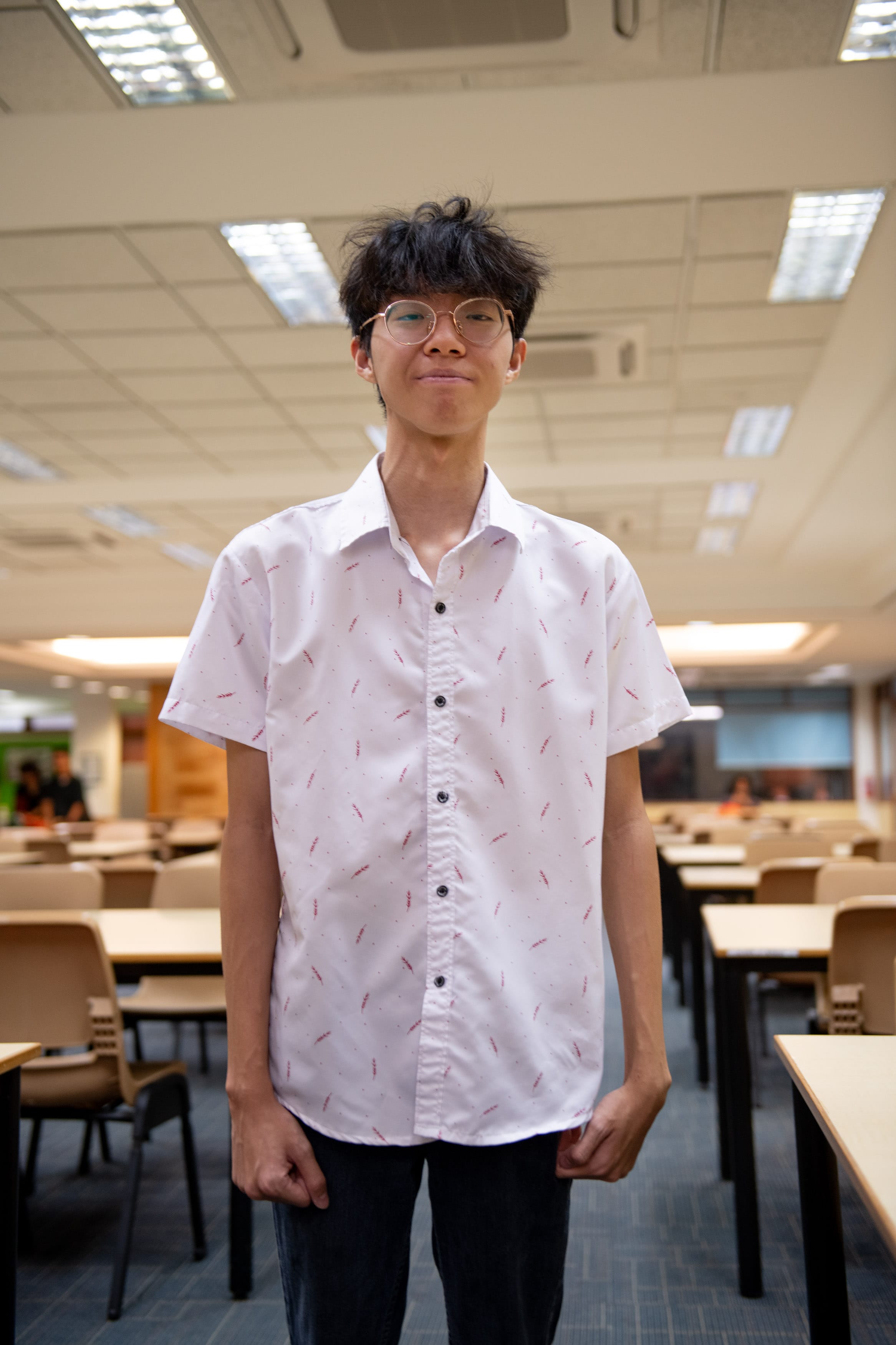 Young man with glasses and dark hair wearing a white patterned shirt, standing in a large room with tables and chairs.