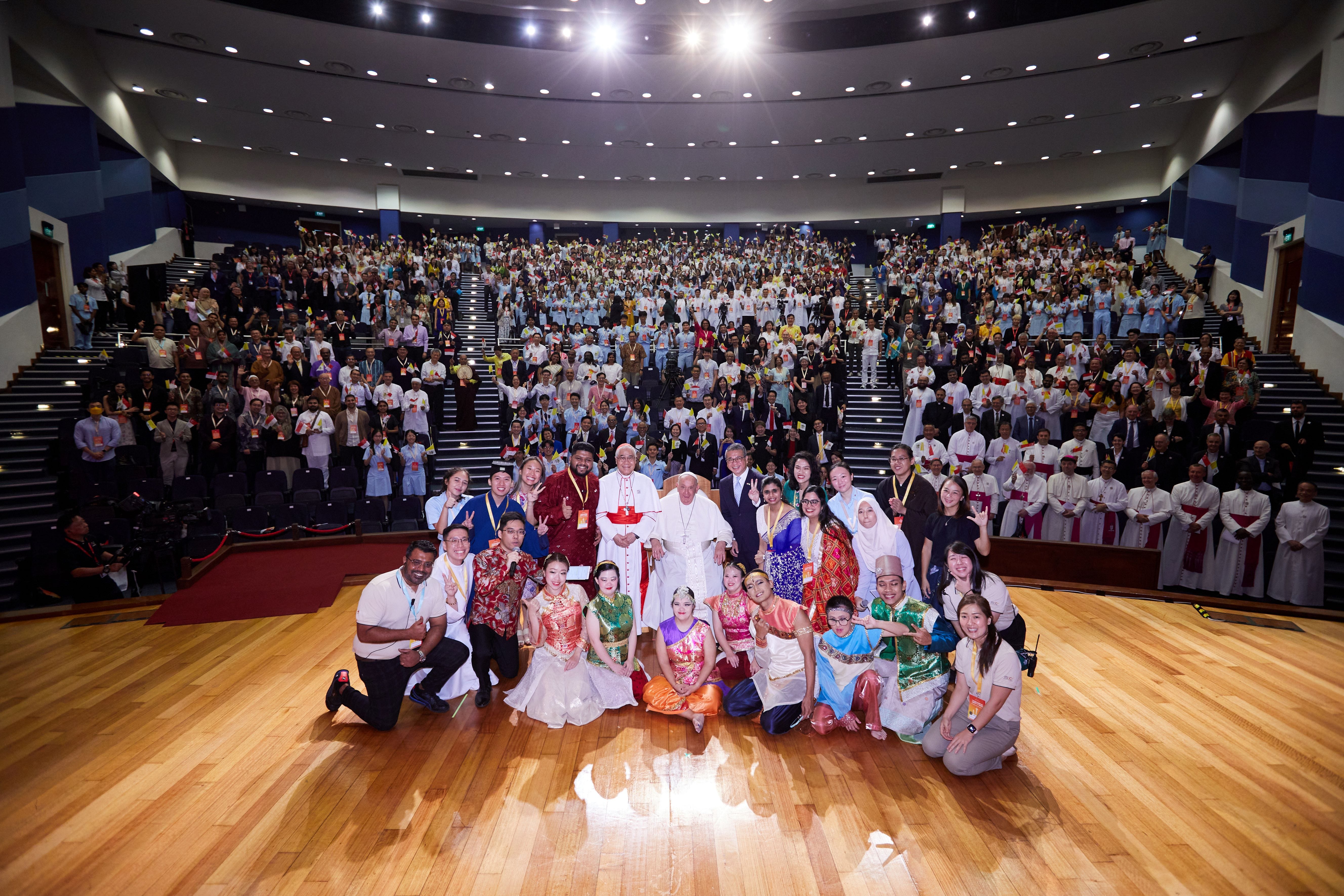 Large group photo in a grand auditorium with hundreds of people in diverse traditional and formal attire posing together.