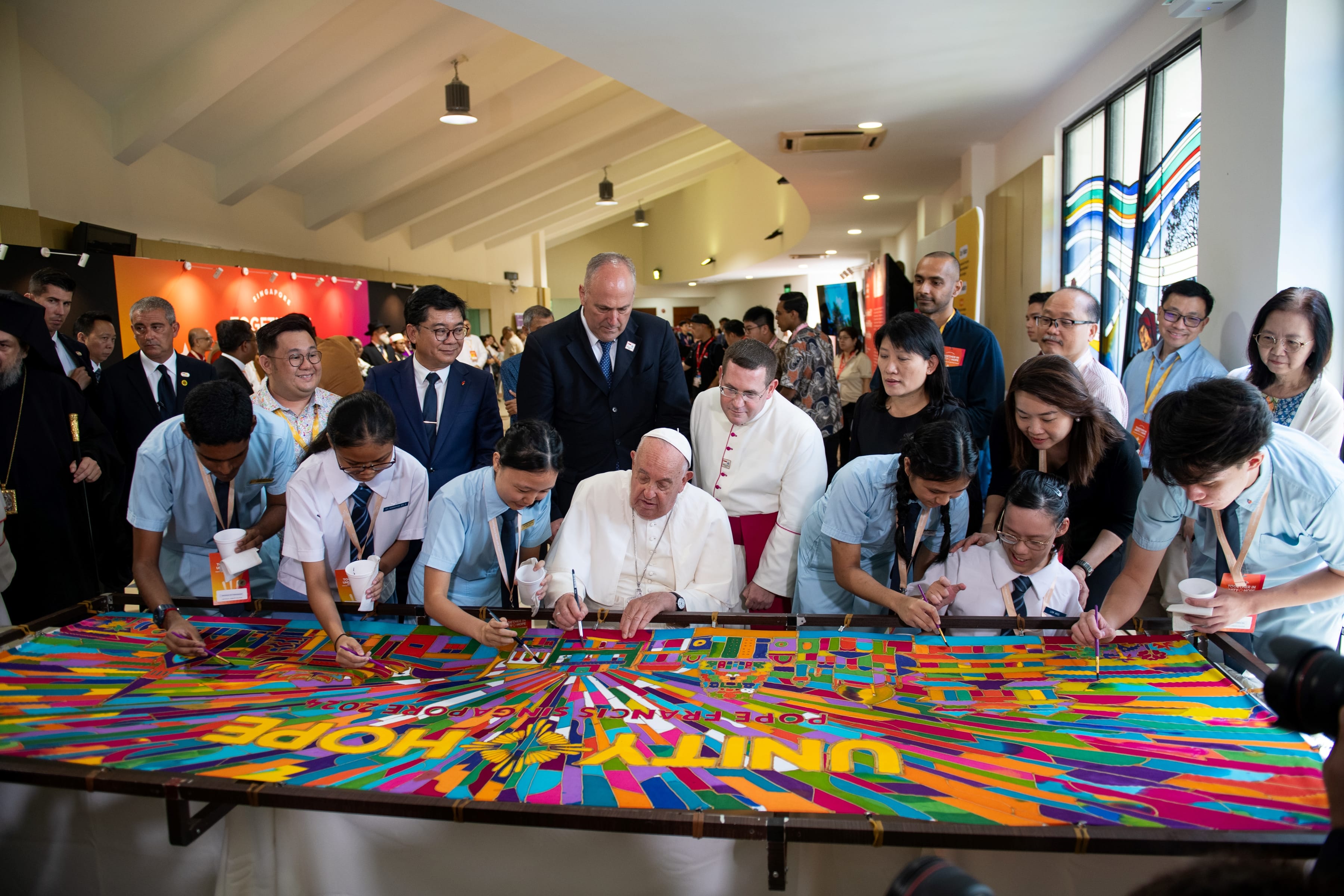 Group gathered as seated man and students paint a large colorful banner on a table in a crowded hall.