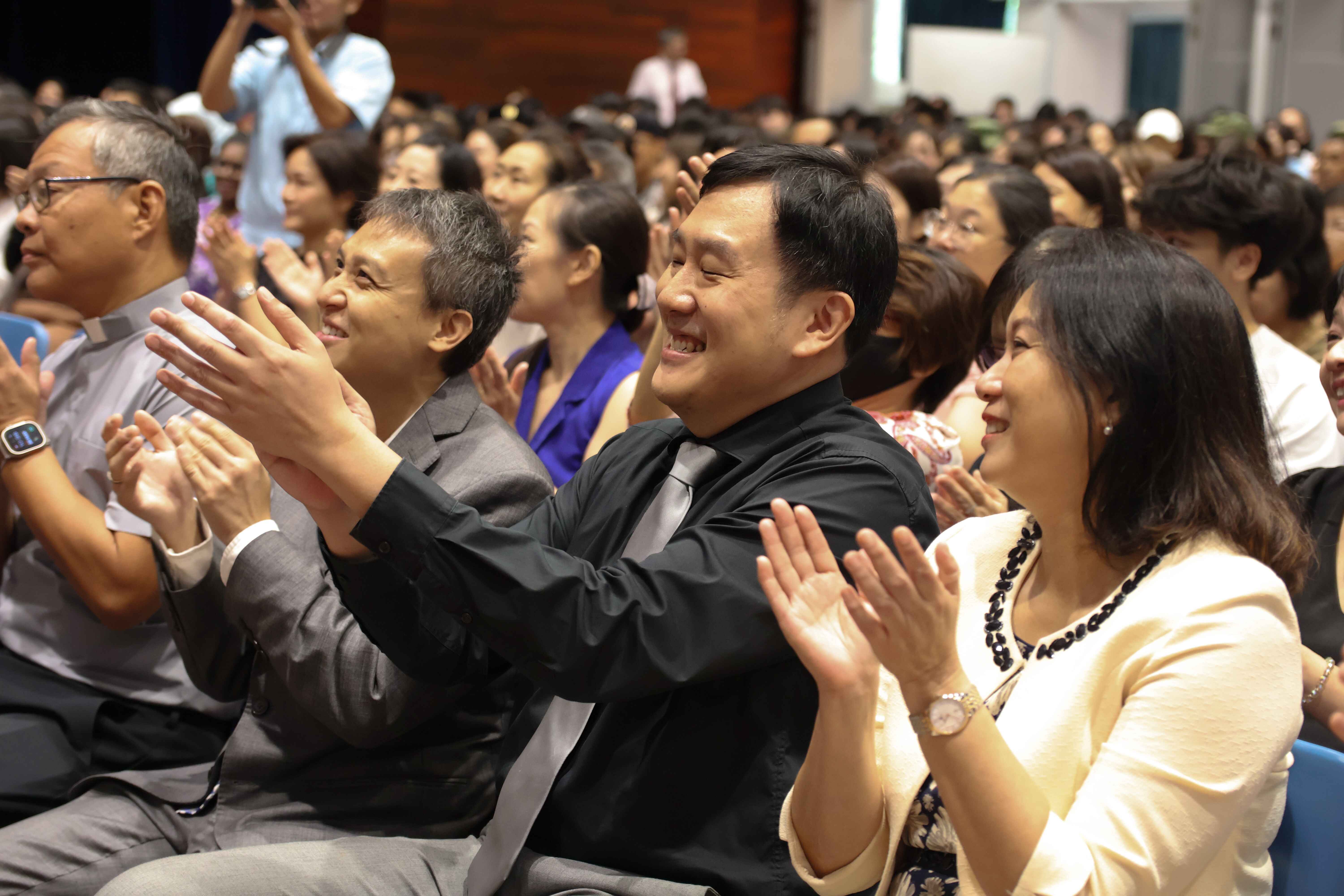 A large seated crowd applauds and smiles at an indoor event, with three people clearly visible in the foreground.