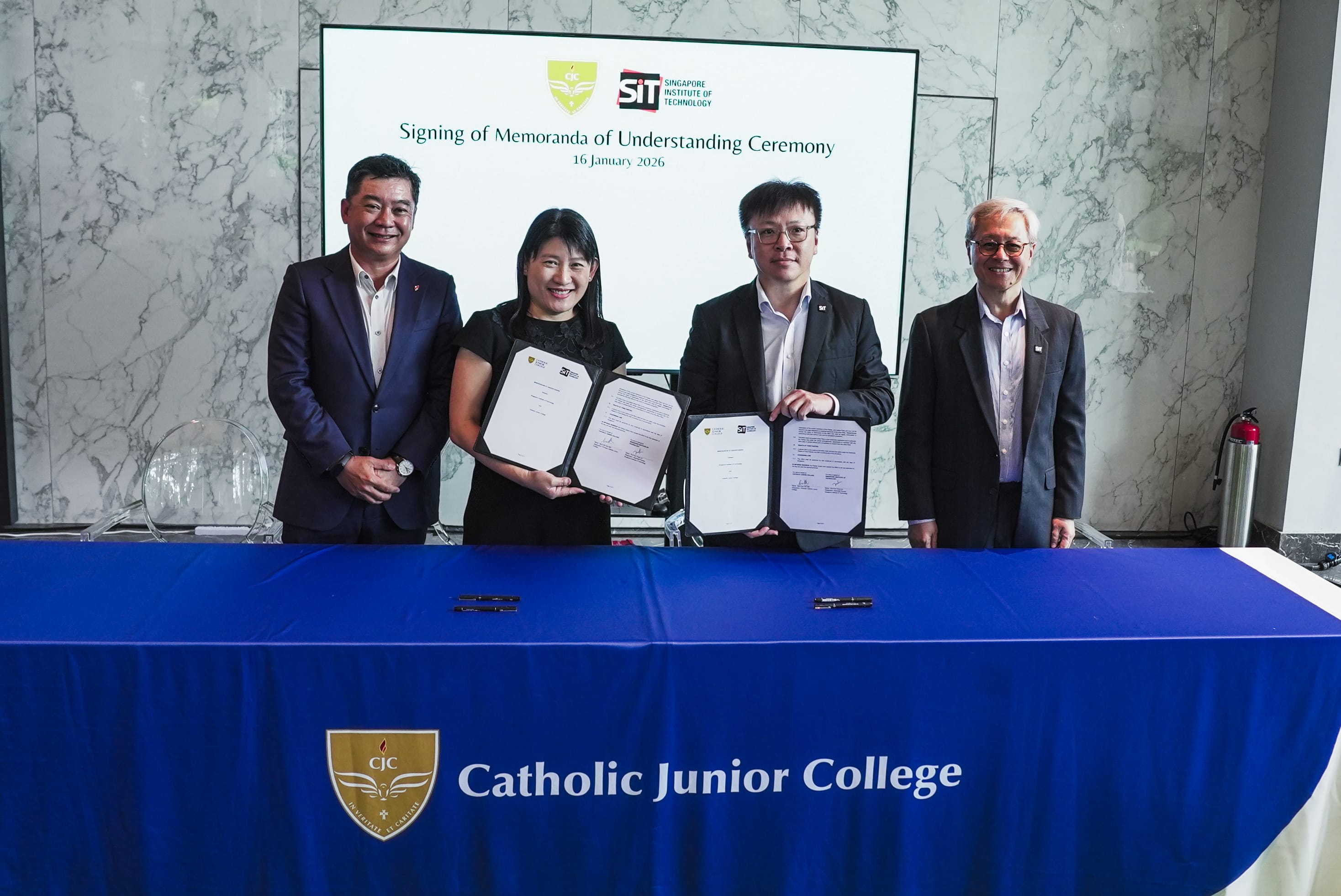 Four people stand at a Catholic Junior College table holding signed MOU documents at a ceremony on 16 January 2026.