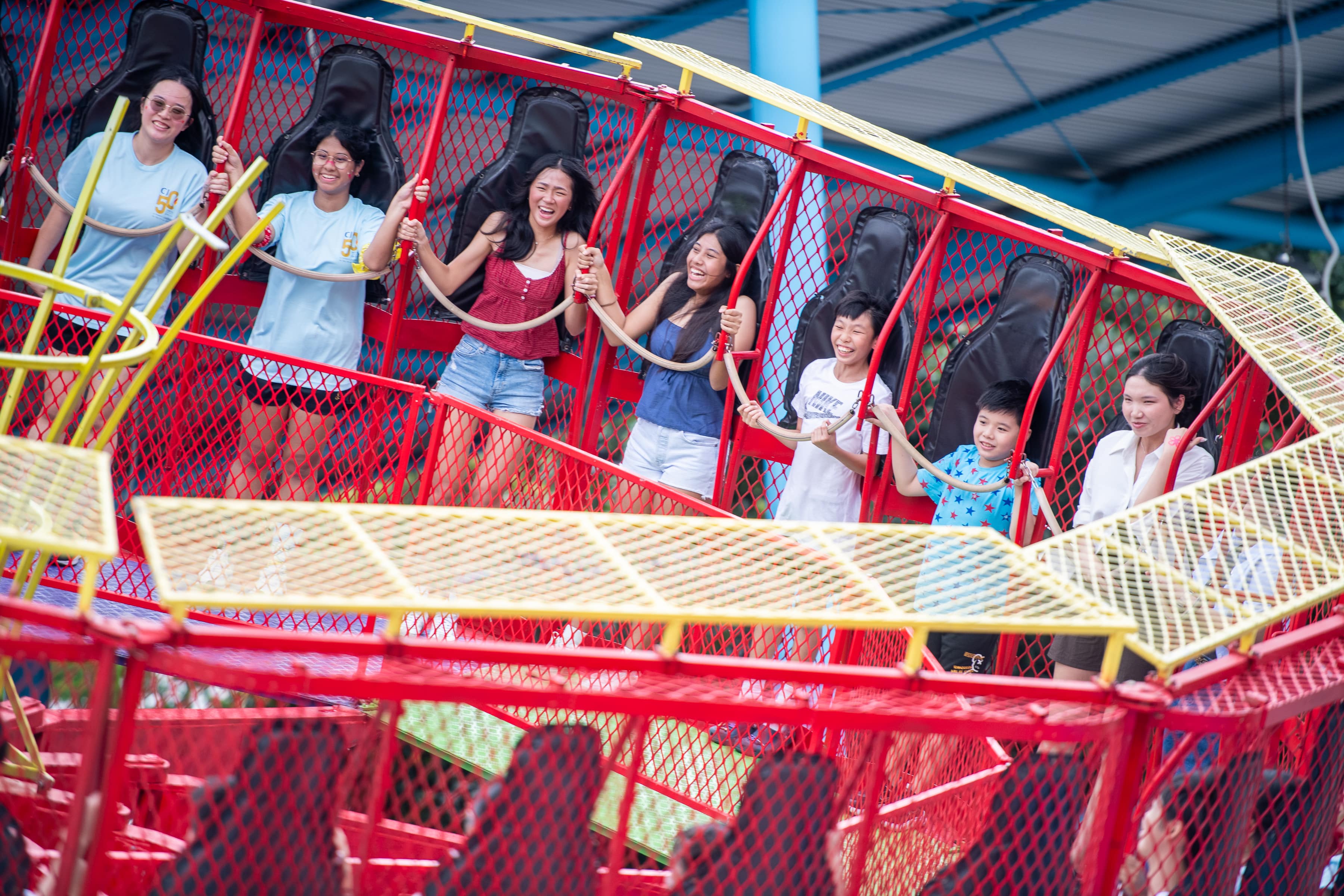 Six laughing people ride a tilting red and yellow amusement park attraction, gripping rope handles.