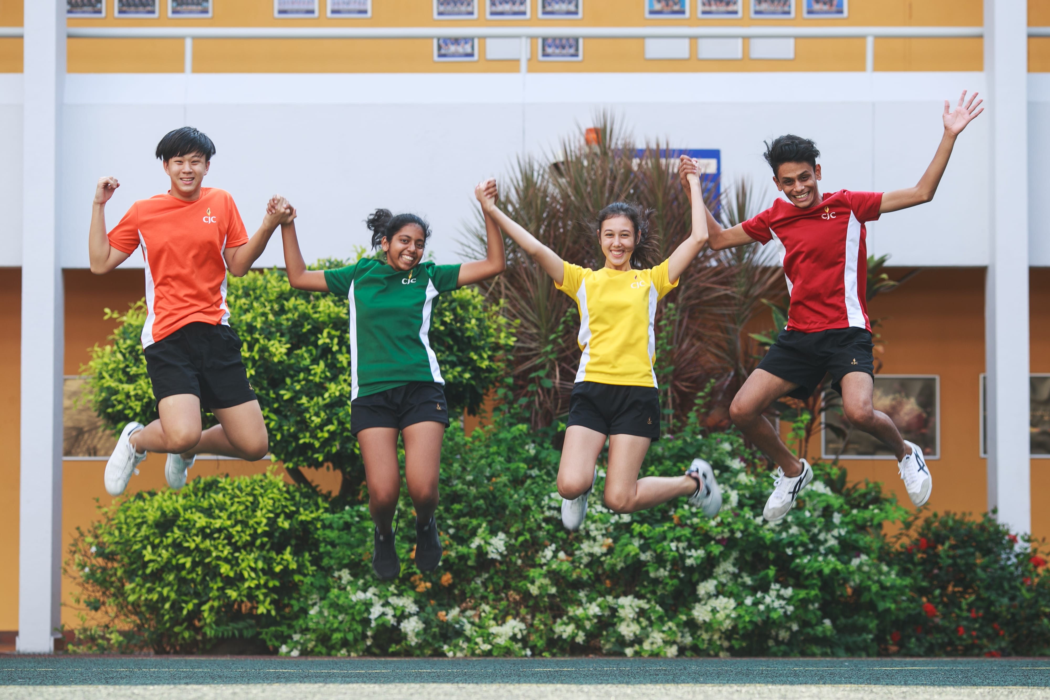 Four students in colorful sports shirts jump holding hands in front of a school building and bushes