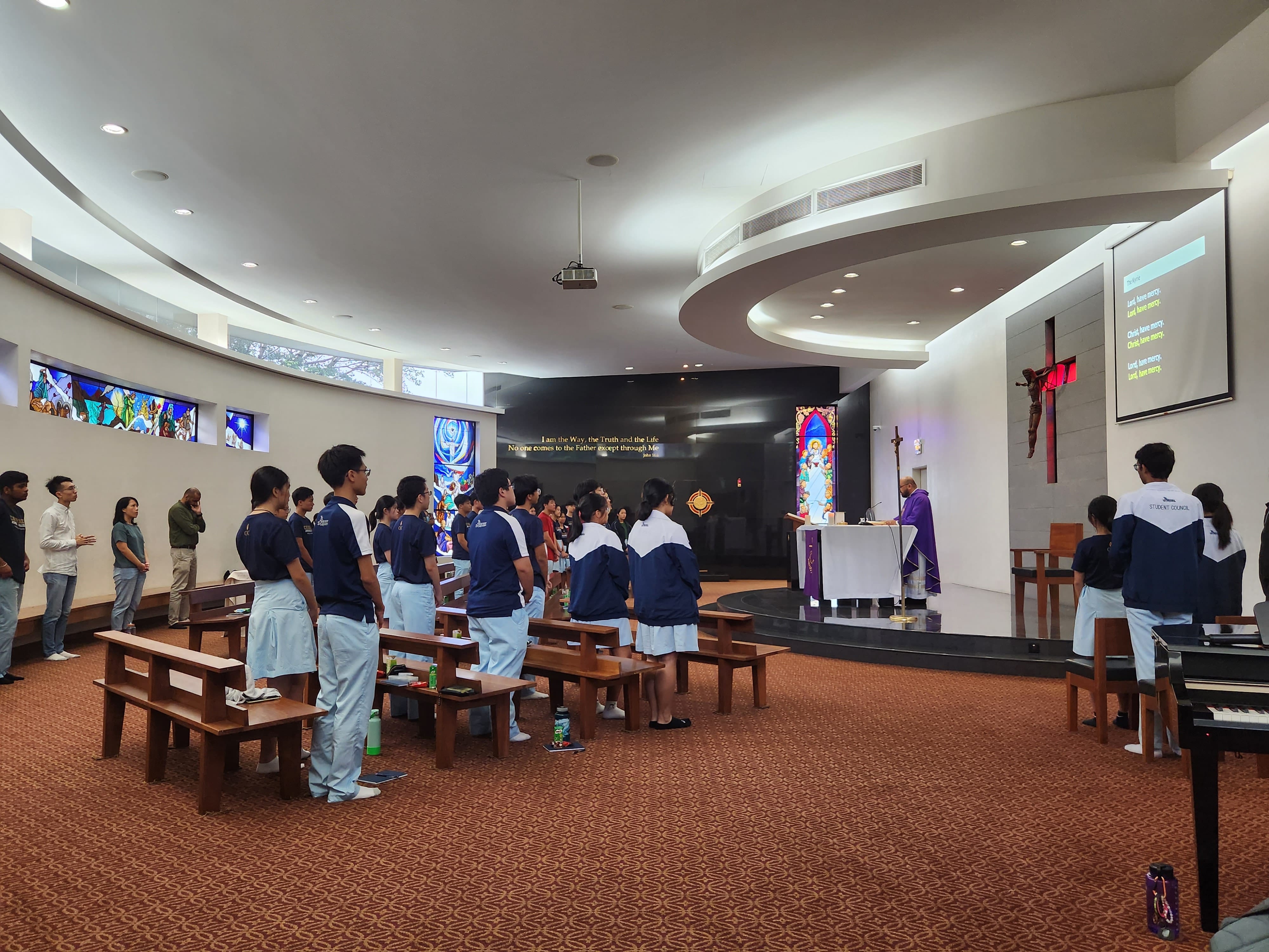 Students in uniforms stand during a Catholic Mass in a modern chapel with stained glass, a cross, and an altar.