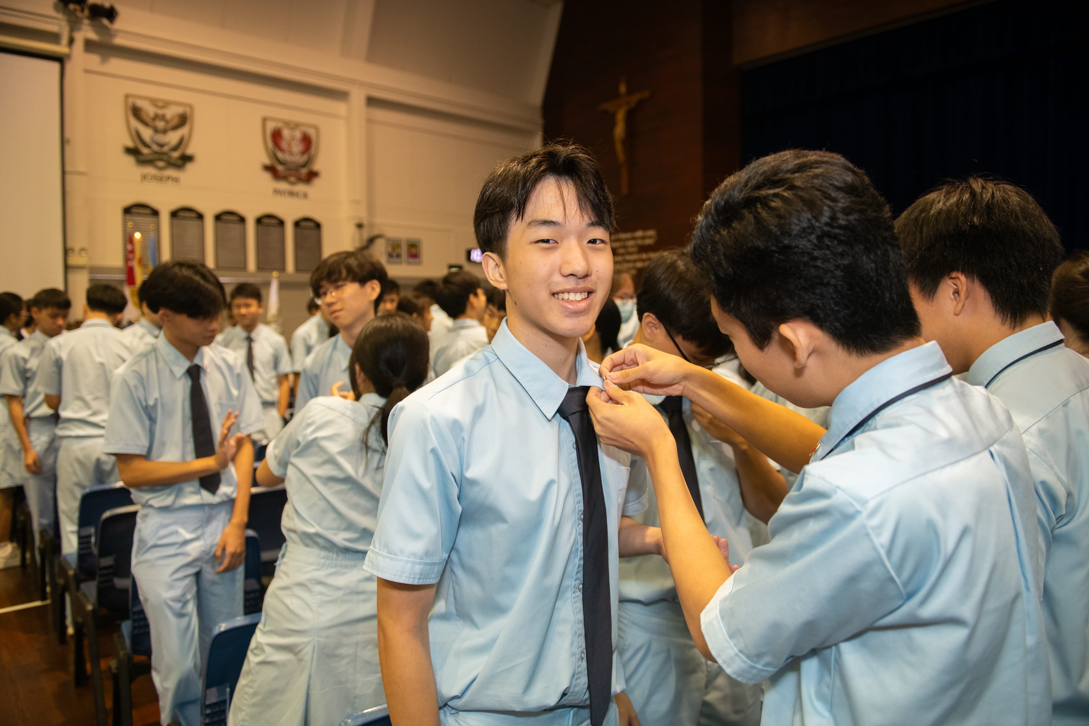 Smiling student in light blue uniform while another student adjusts his tie in a crowded school hall.
