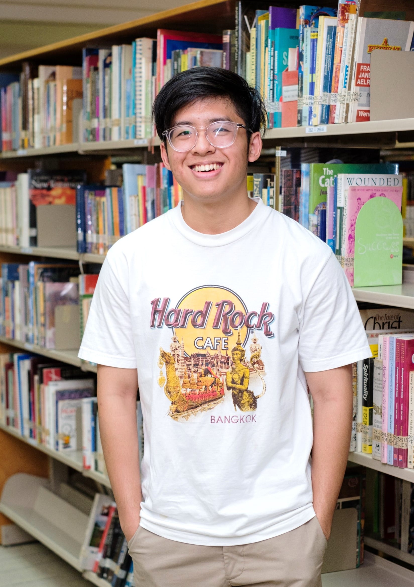 Young man with glasses smiling in a library, wearing a white Hard Rock Cafe Bangkok t-shirt.