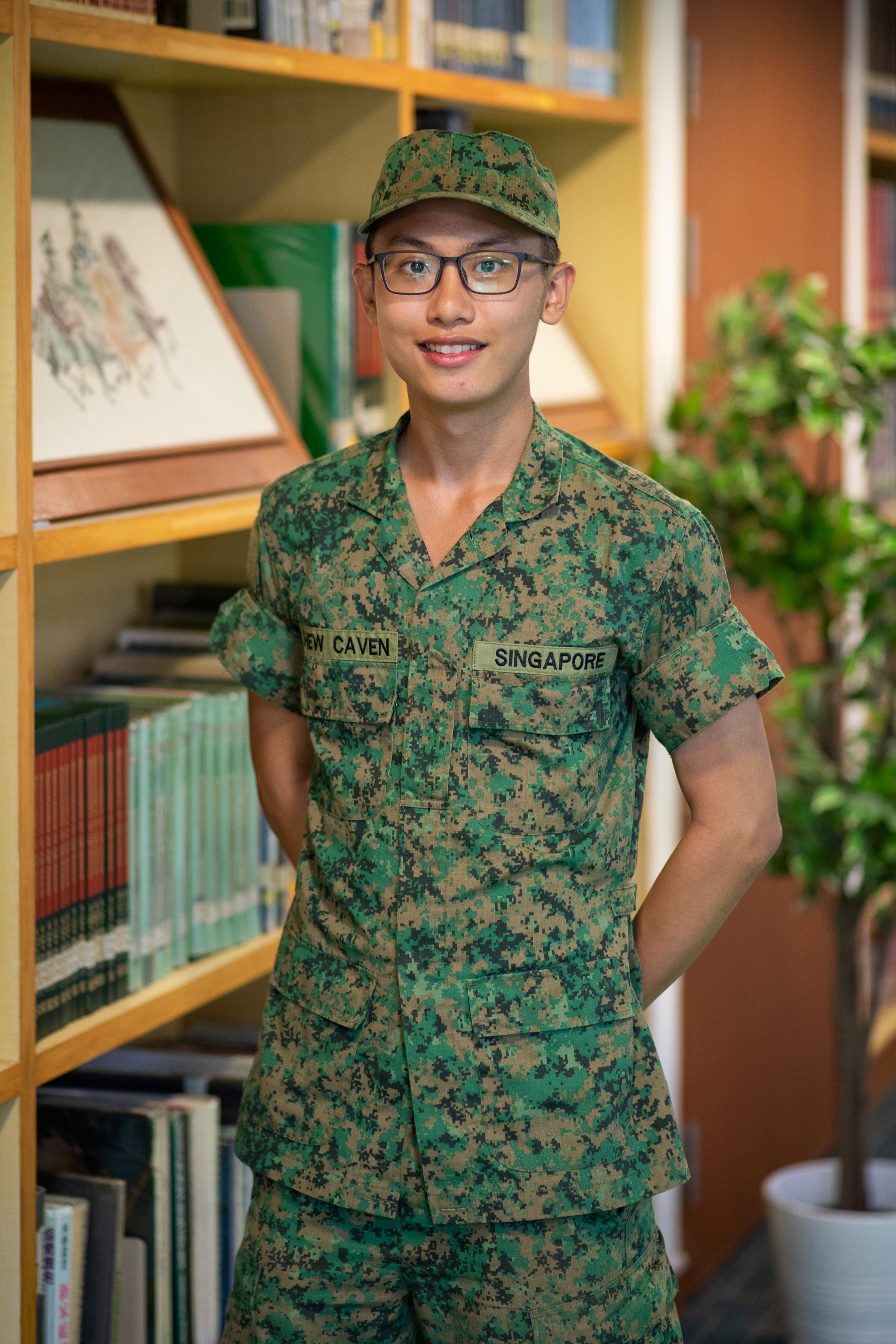 Young man in Singapore military camouflage uniform and cap, wearing glasses, smiling in front of a bookshelf.