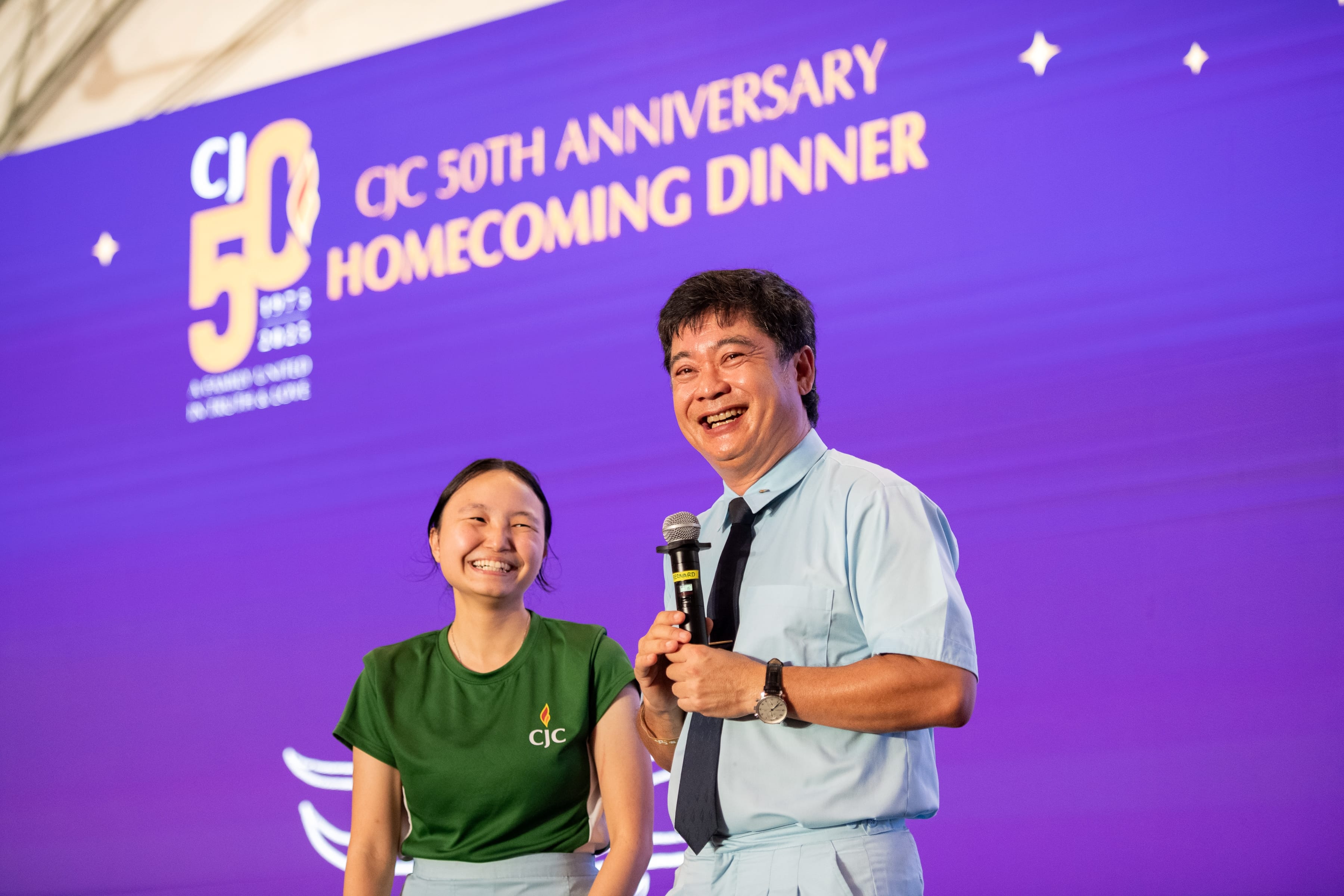 Smiling man with microphone stands beside smiling woman on stage with purple 50th anniversary backdrop