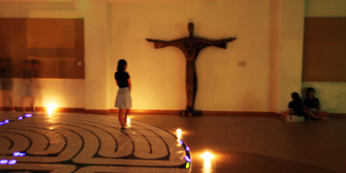 A person stands on a floor labyrinth with candles, facing a wooden crucifix on a dimly lit chapel wall.