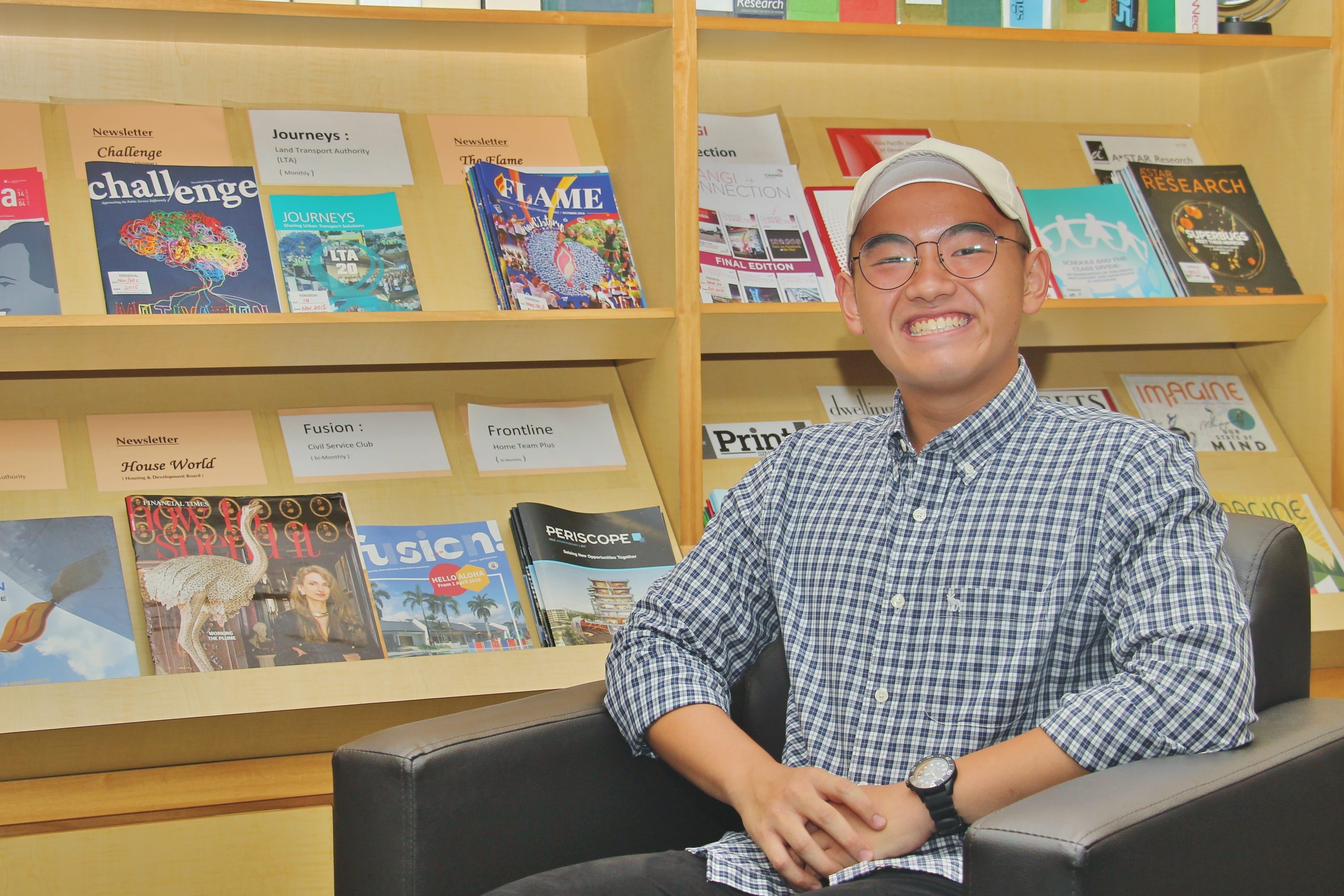 Smiling person in glasses and white cap sits in a black chair before shelves of magazines and newsletters.