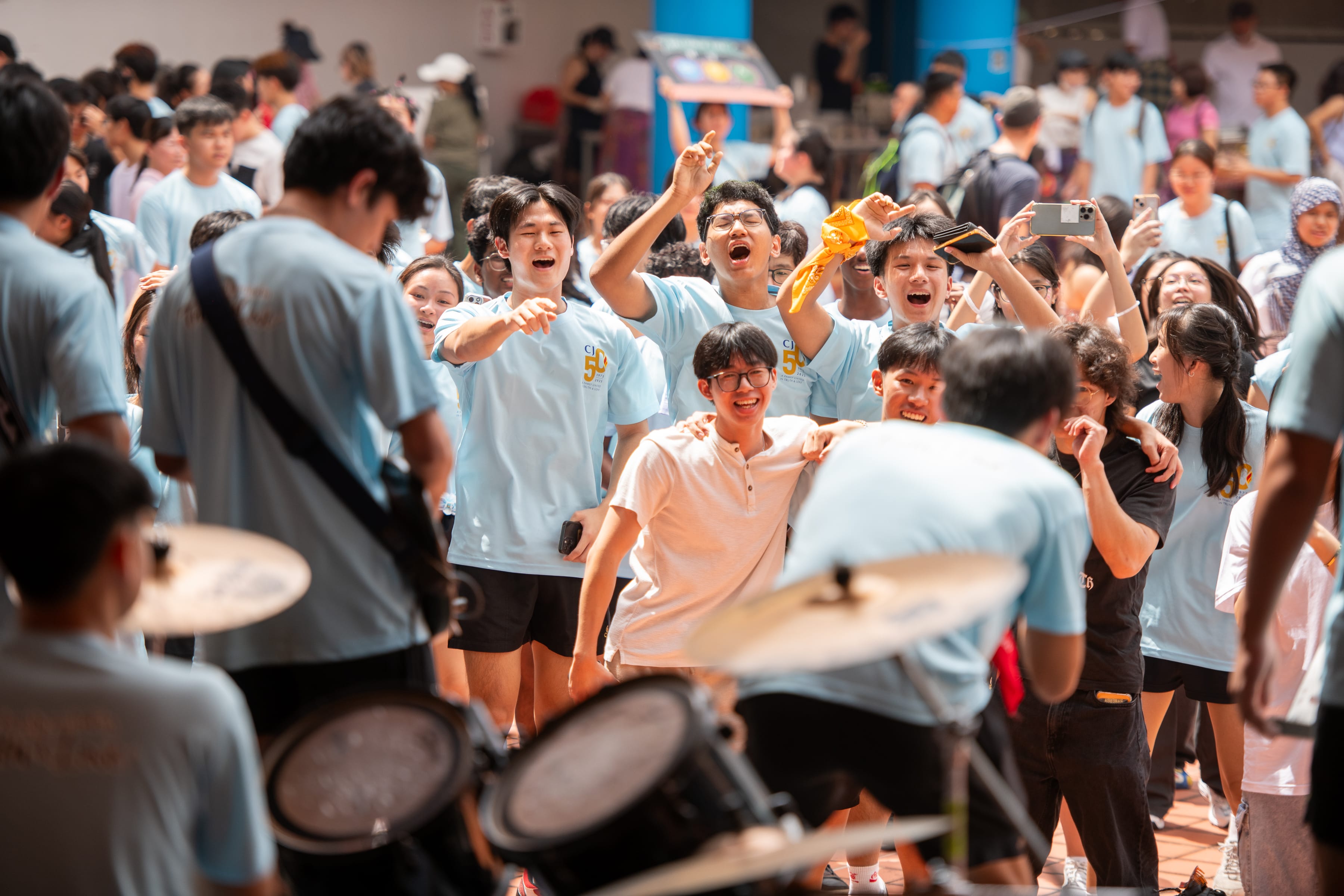 Cheering crowd in light blue shirts watches a band; some raise hands and phones while drums blur in front.