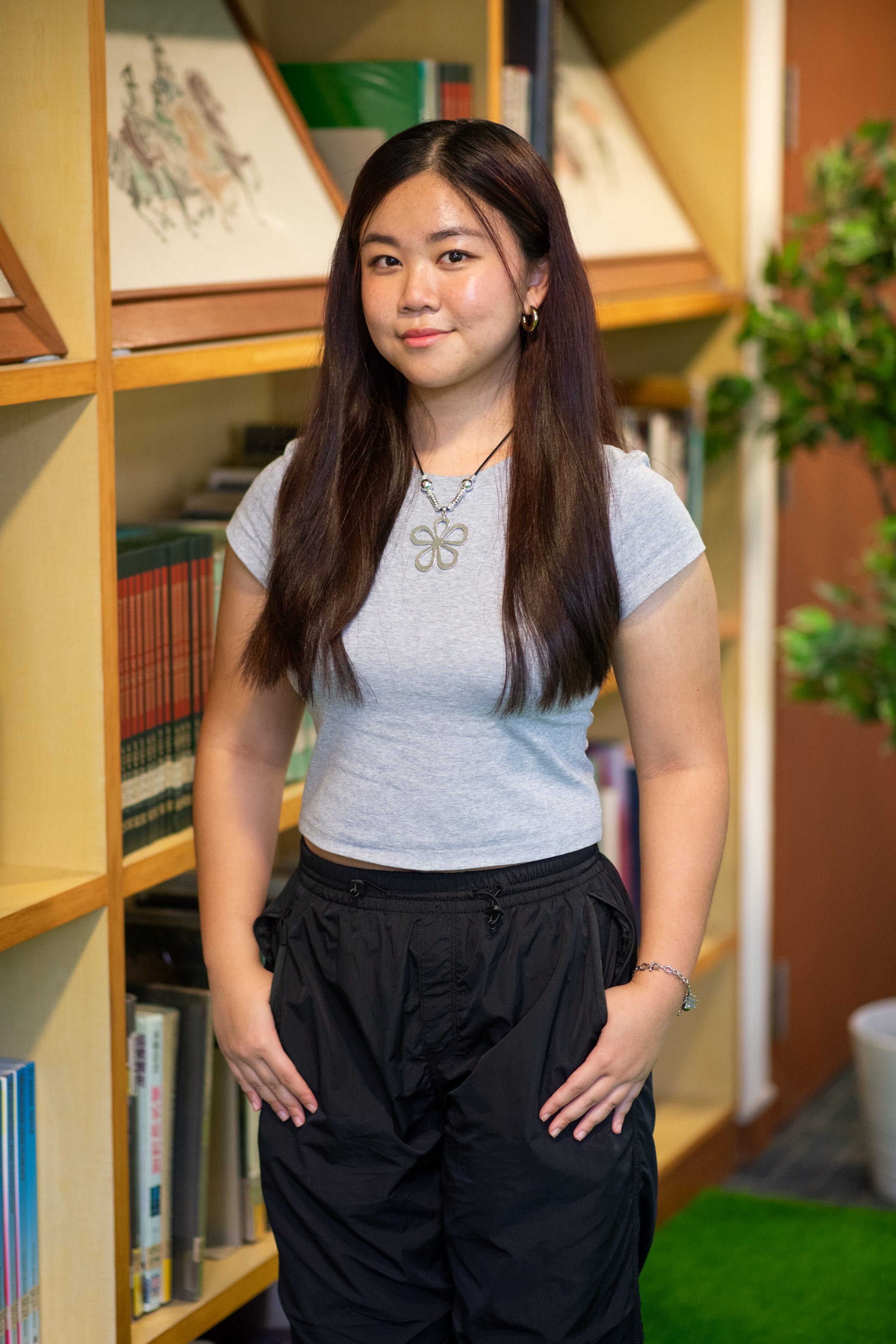 Young woman with long dark hair, gray crop top, black pants, and a flower necklace, standing in front of wooden bookshelves.