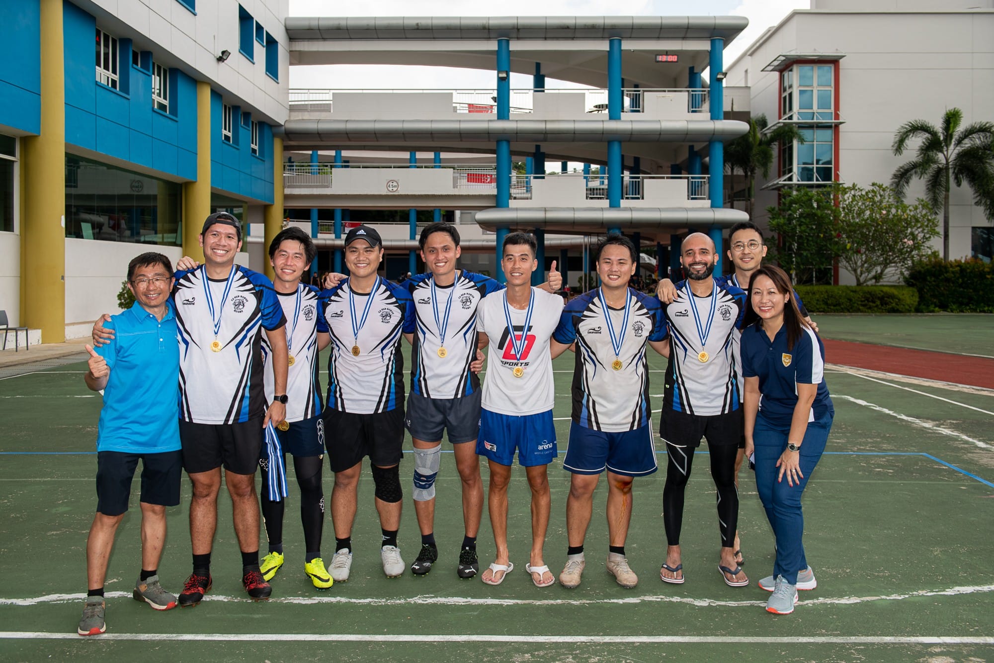 Ten athletes in blue and white jerseys pose with gold medals on an outdoor court beside a modern building.