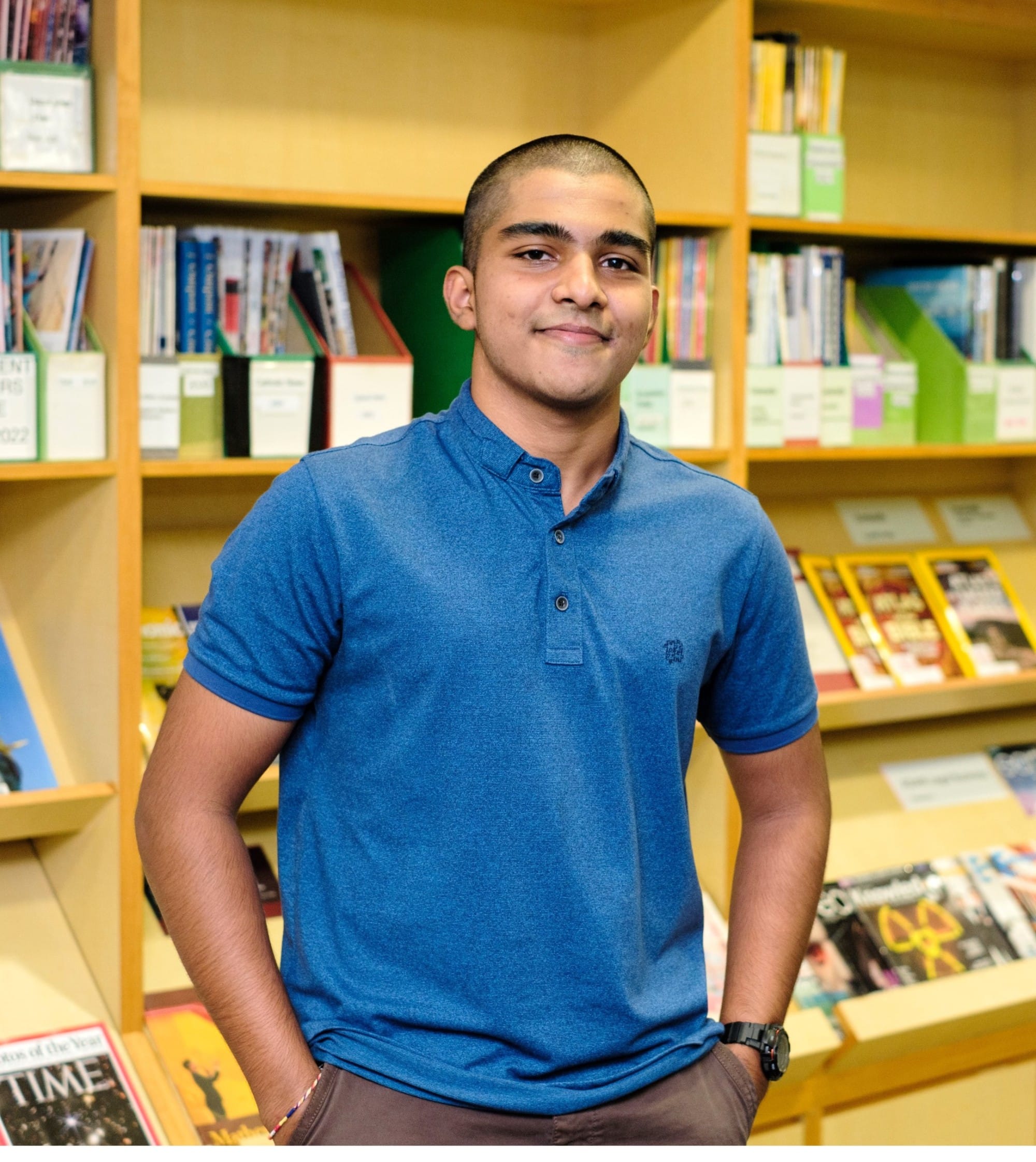 Young man in a blue polo shirt smiling in front of wooden shelves filled with books and magazines.