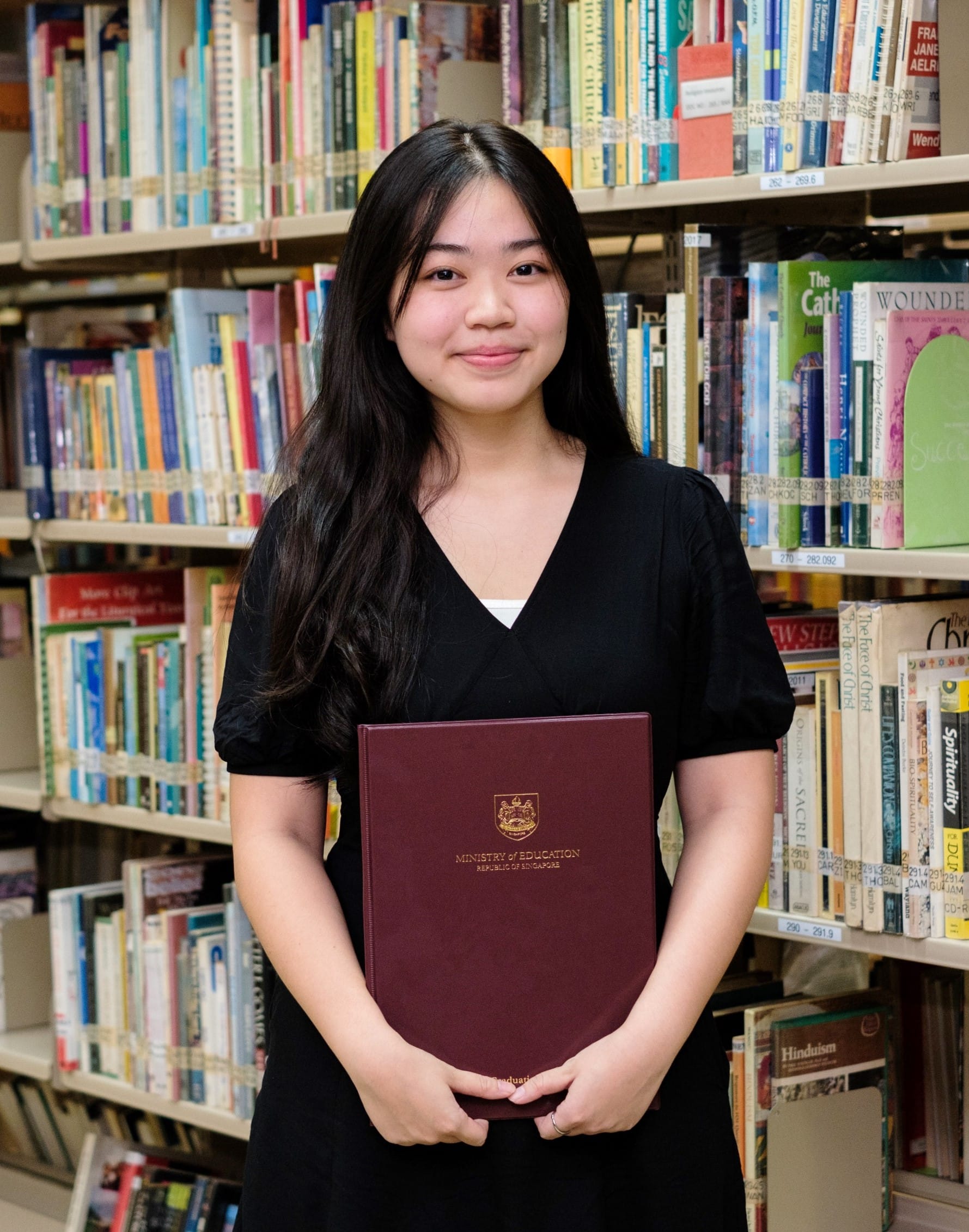 Young woman in black dress holding a red Ministry of Education Singapore certificate folder, smiling in a library.