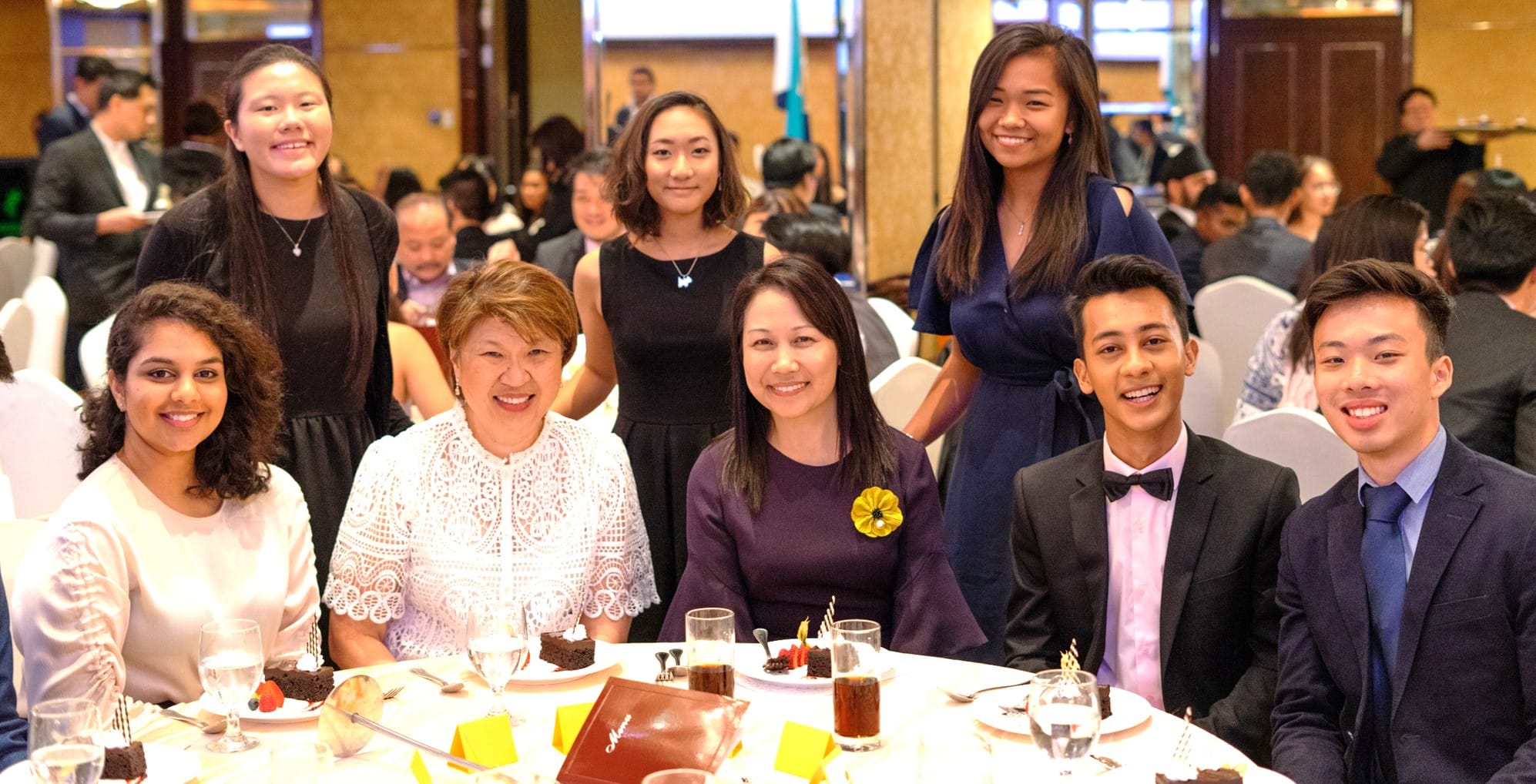 Eight formally dressed people smile at a banquet table with dessert plates, drinks, and a menu in a crowded hall.