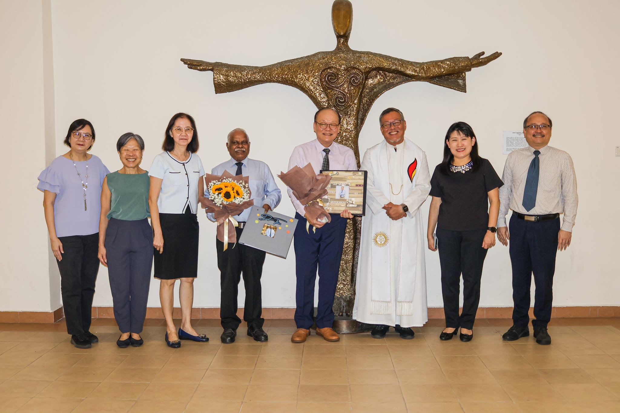 Seven people pose indoors before a bronze cross sculpture; two hold flowers and a framed gift, one wears white clergy robes.