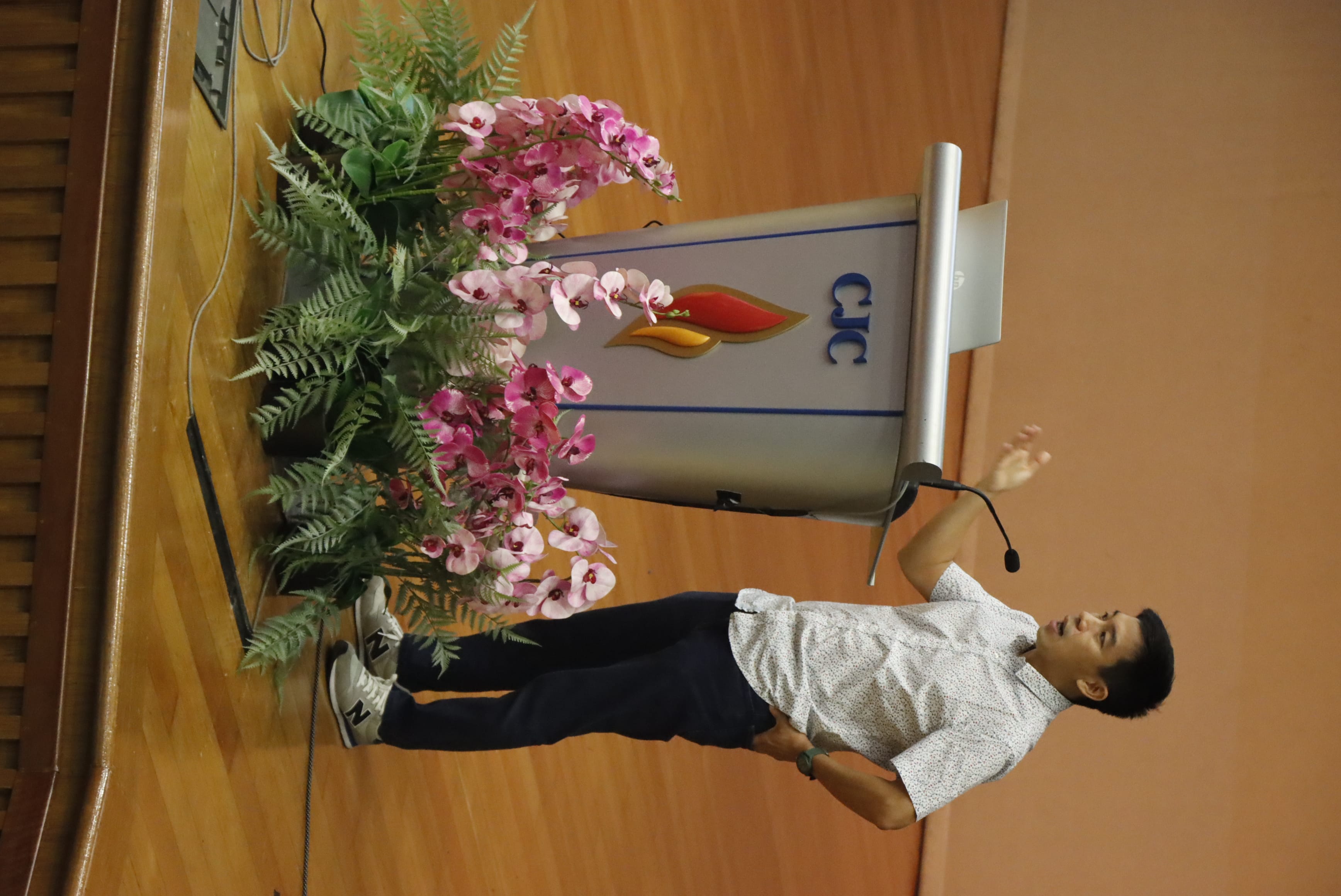 Man in white shirt speaking at a CJC podium with pink orchids, gesturing with one hand on a wooden stage.