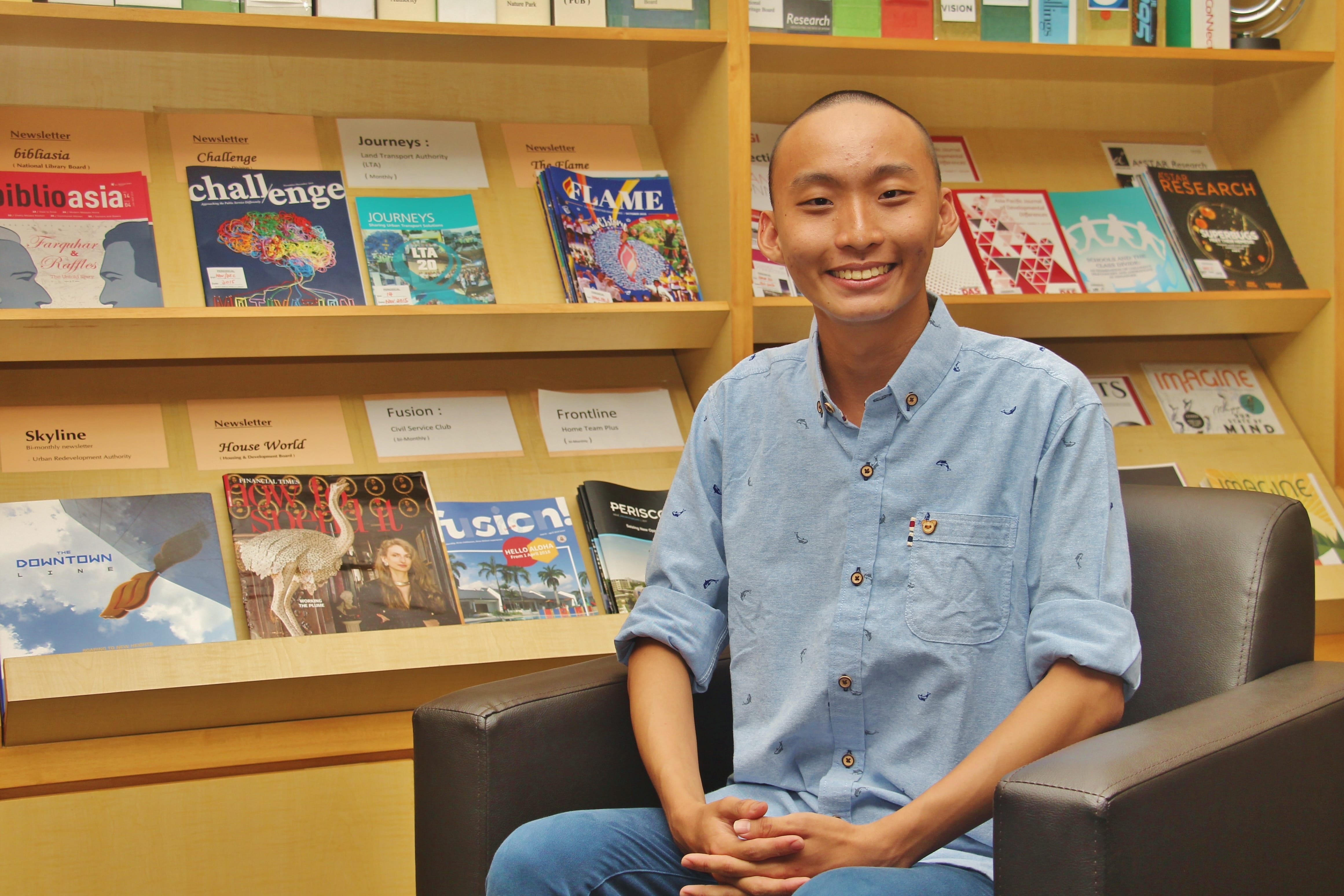 Smiling person in blue shirt sits in an armchair in front of wooden shelves filled with magazines.