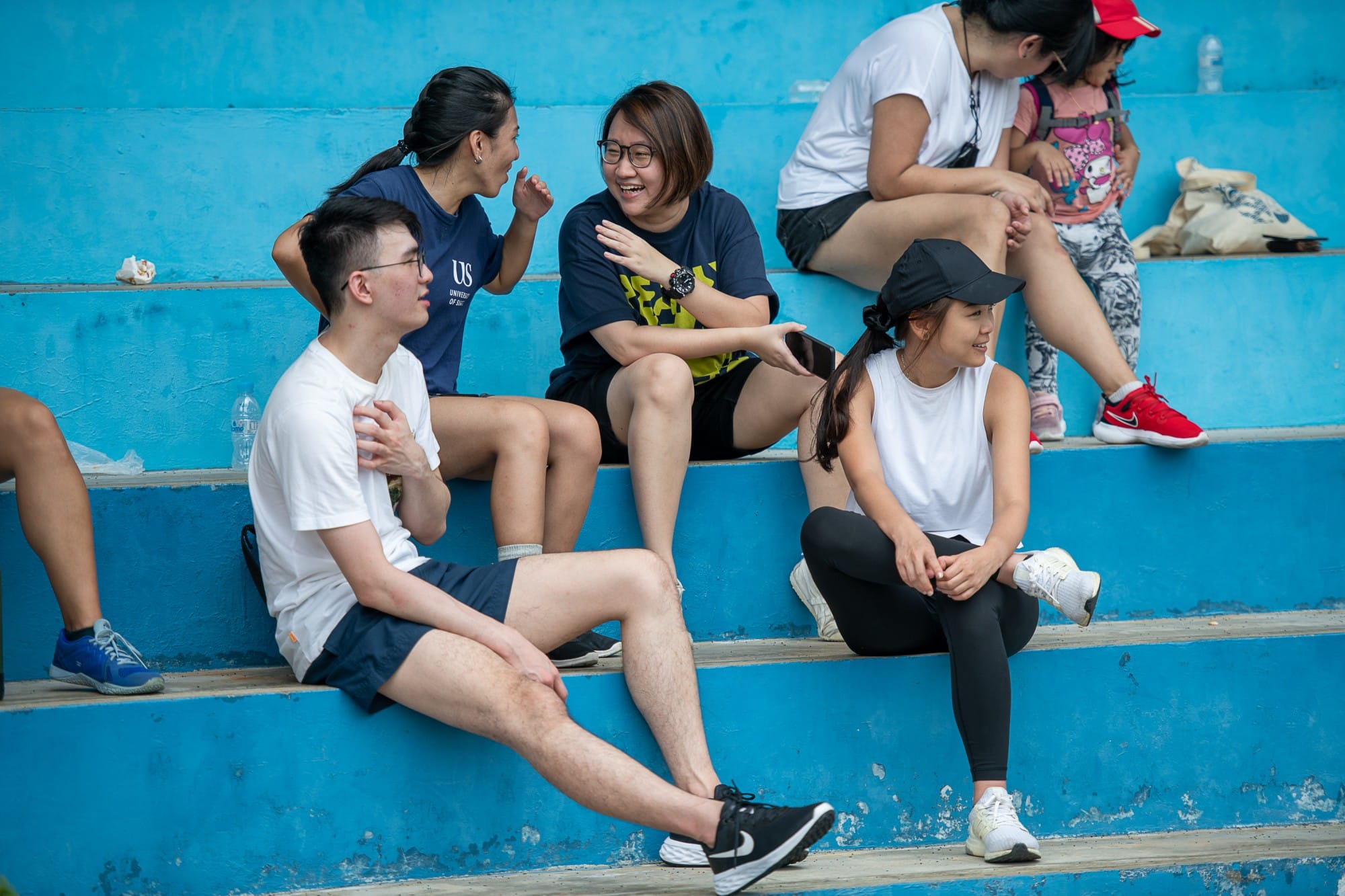 Several young people in athletic wear sit and chat on blue painted bleacher steps outdoors.
