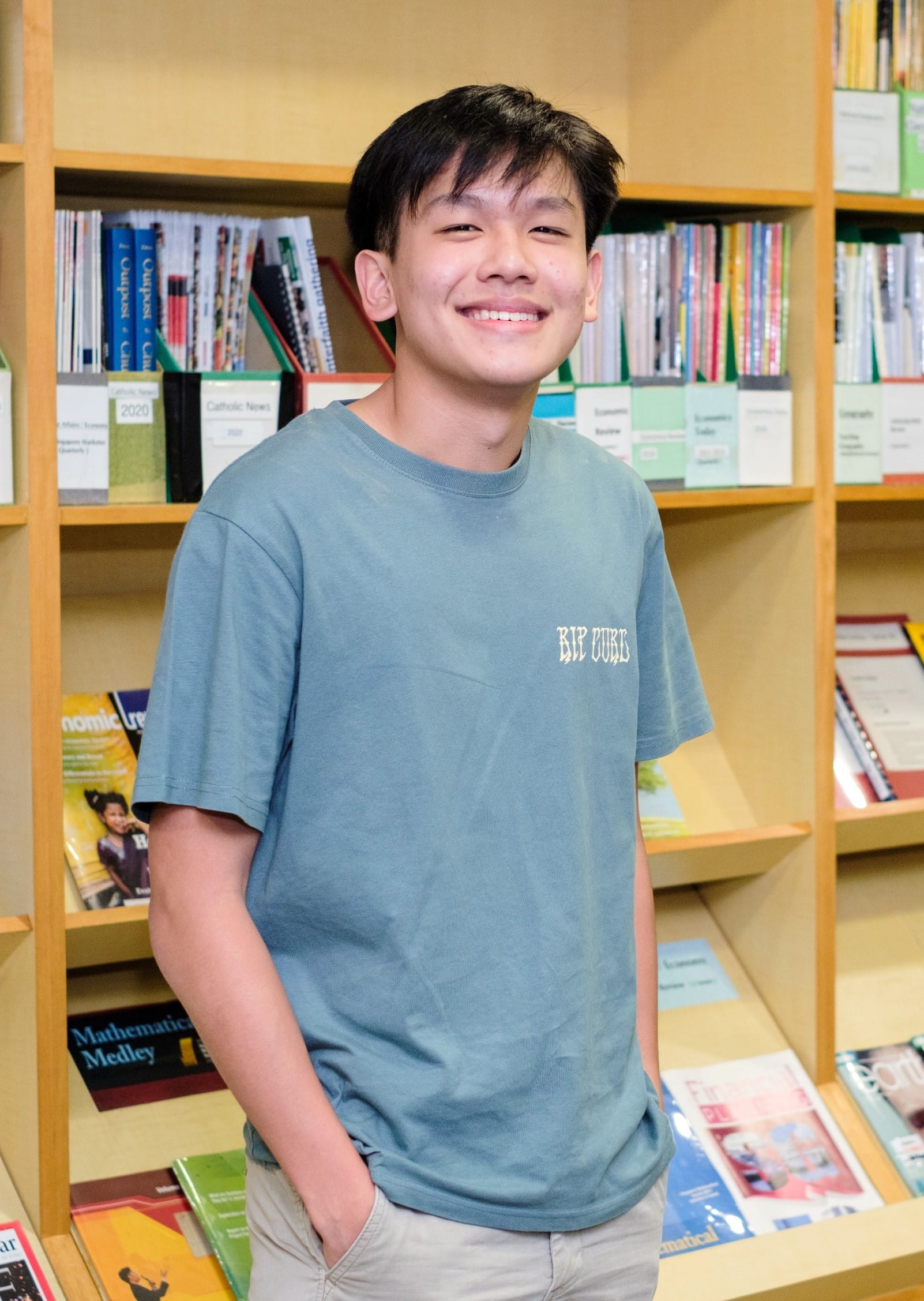 Young man smiling in a blue Rip Curl t-shirt, standing in front of wooden shelves filled with books.