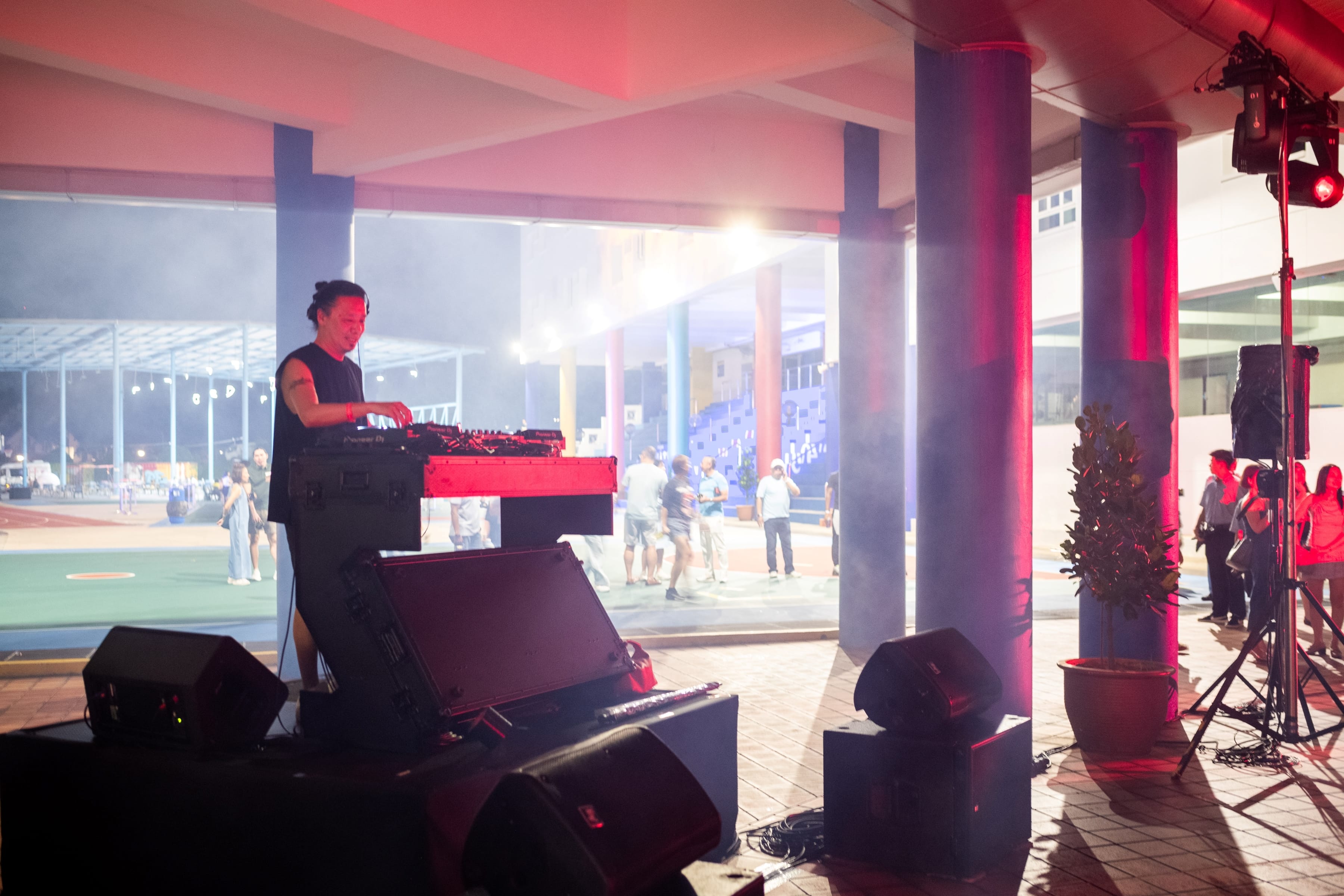 DJ at a mixing table on a small stage with speakers, red lighting, and people in the background.