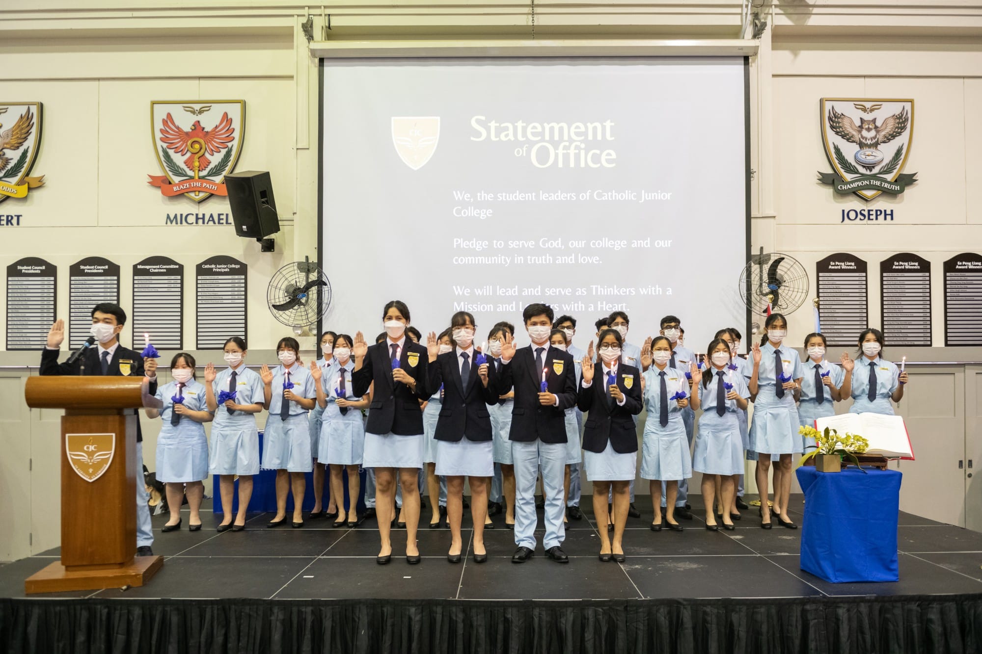 Students in uniform raise their hands on stage during a Catholic Junior College Statement of Office ceremony.