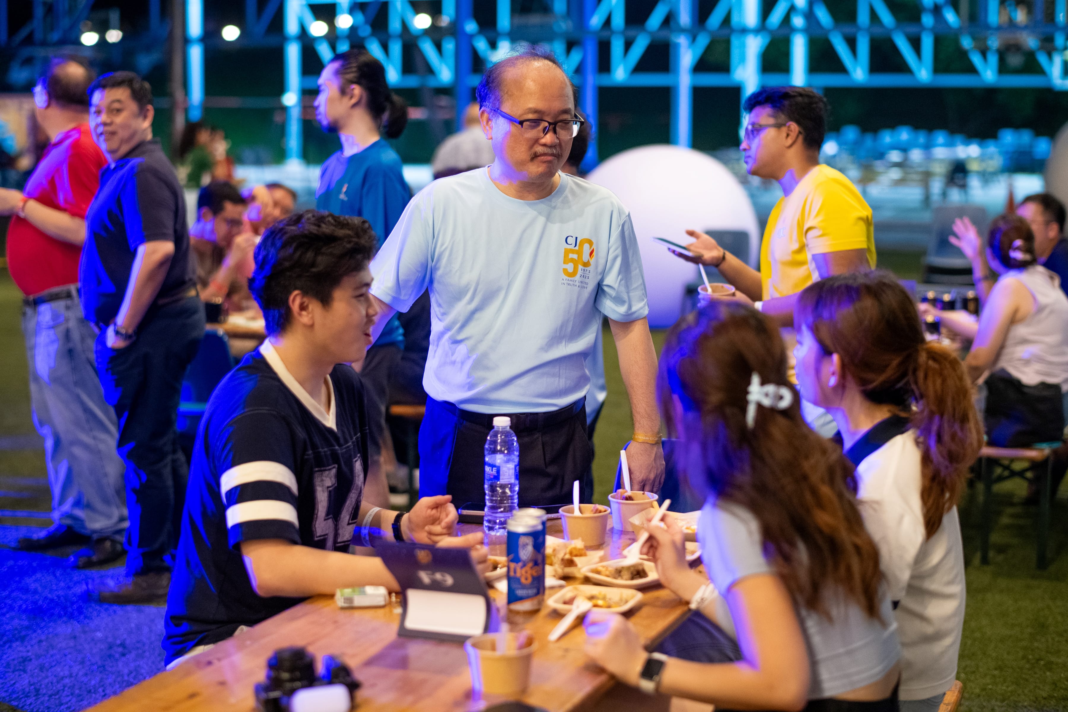 Man in light blue shirt stands by table as group eats and chats at an outdoor night gathering with blue lights.