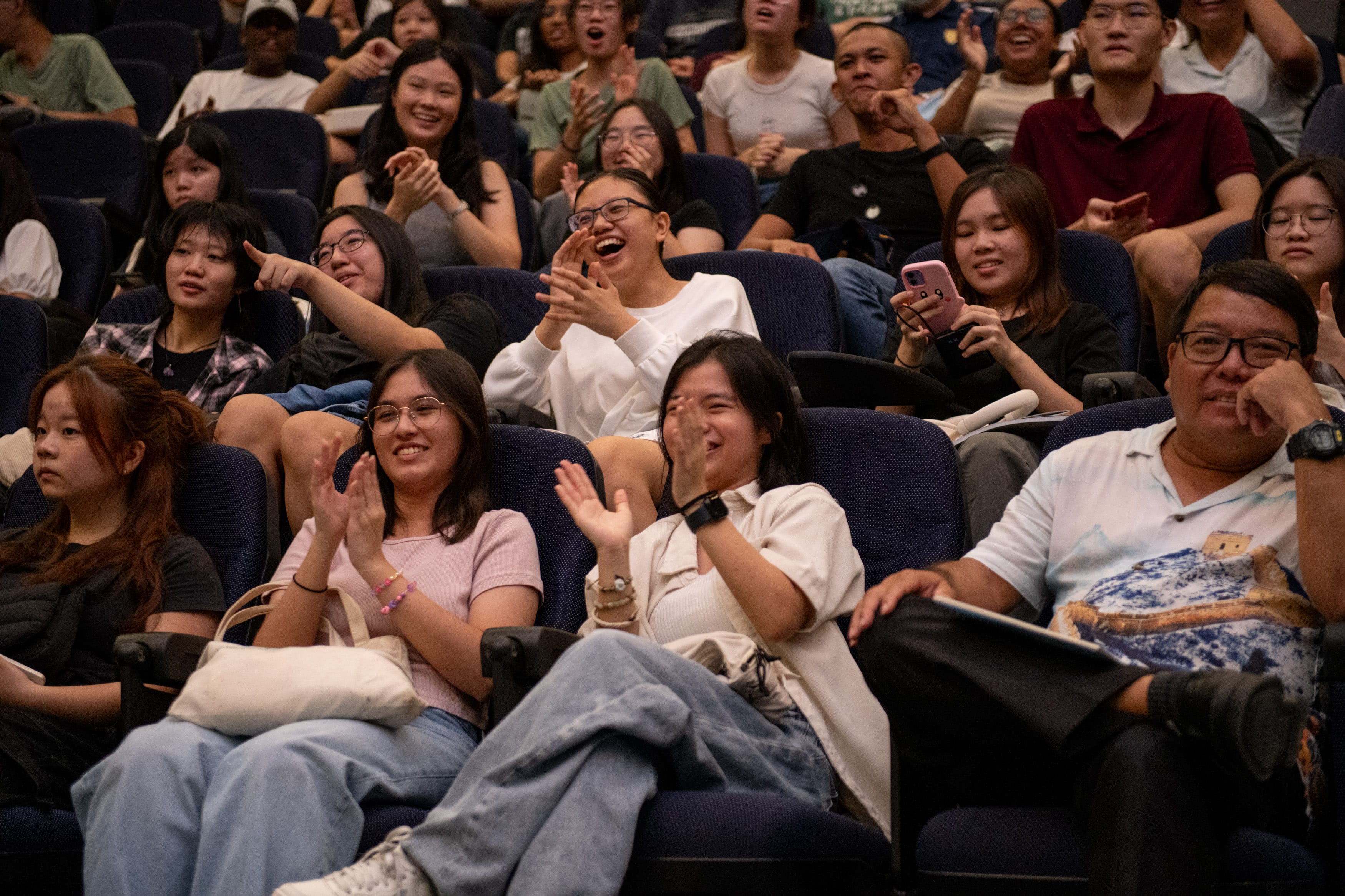 A large crowd of young people sit in auditorium seats, laughing and clapping with joyful expressions.