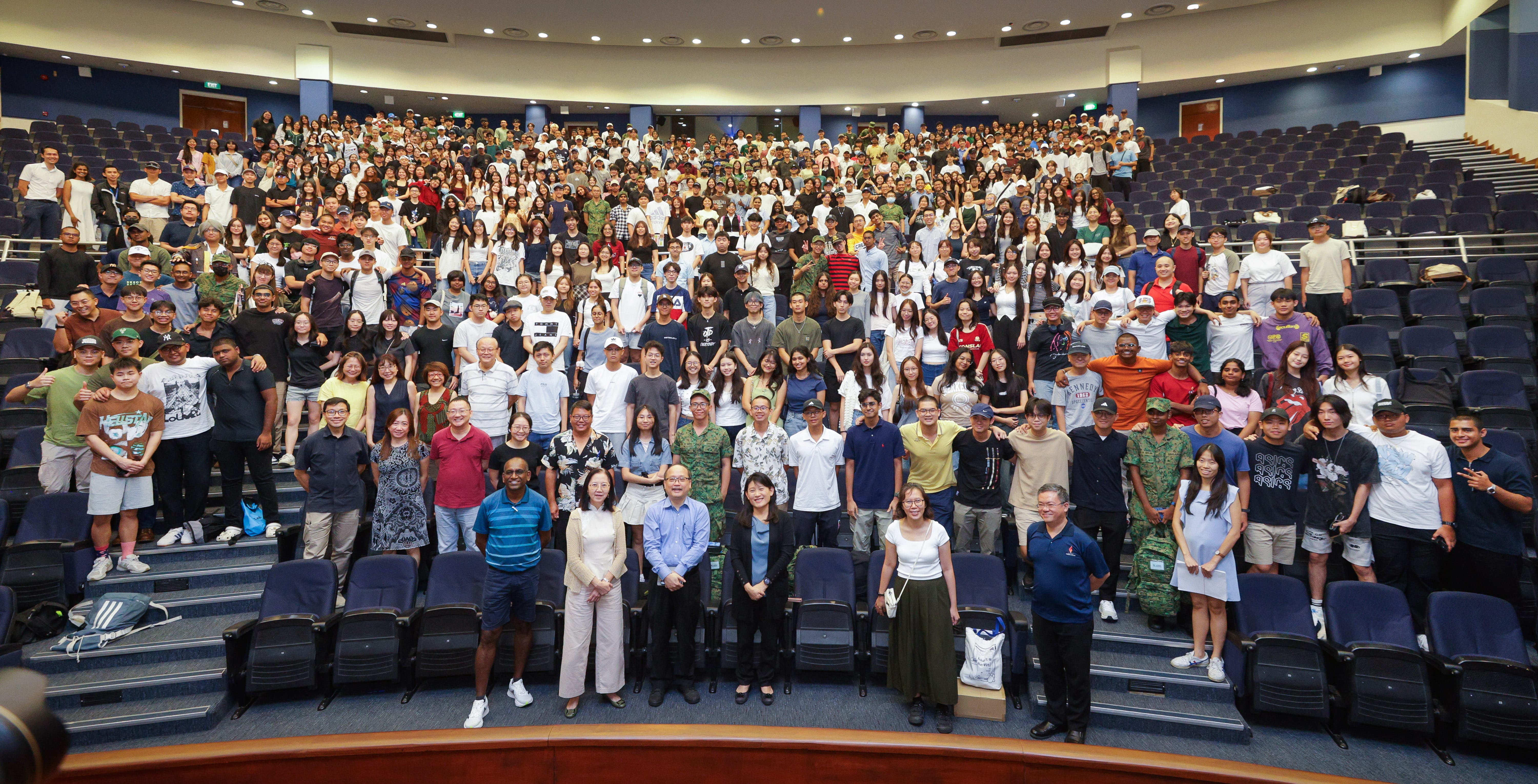 Large group of hundreds of people posing for a photo inside a large auditorium with tiered blue seats.
