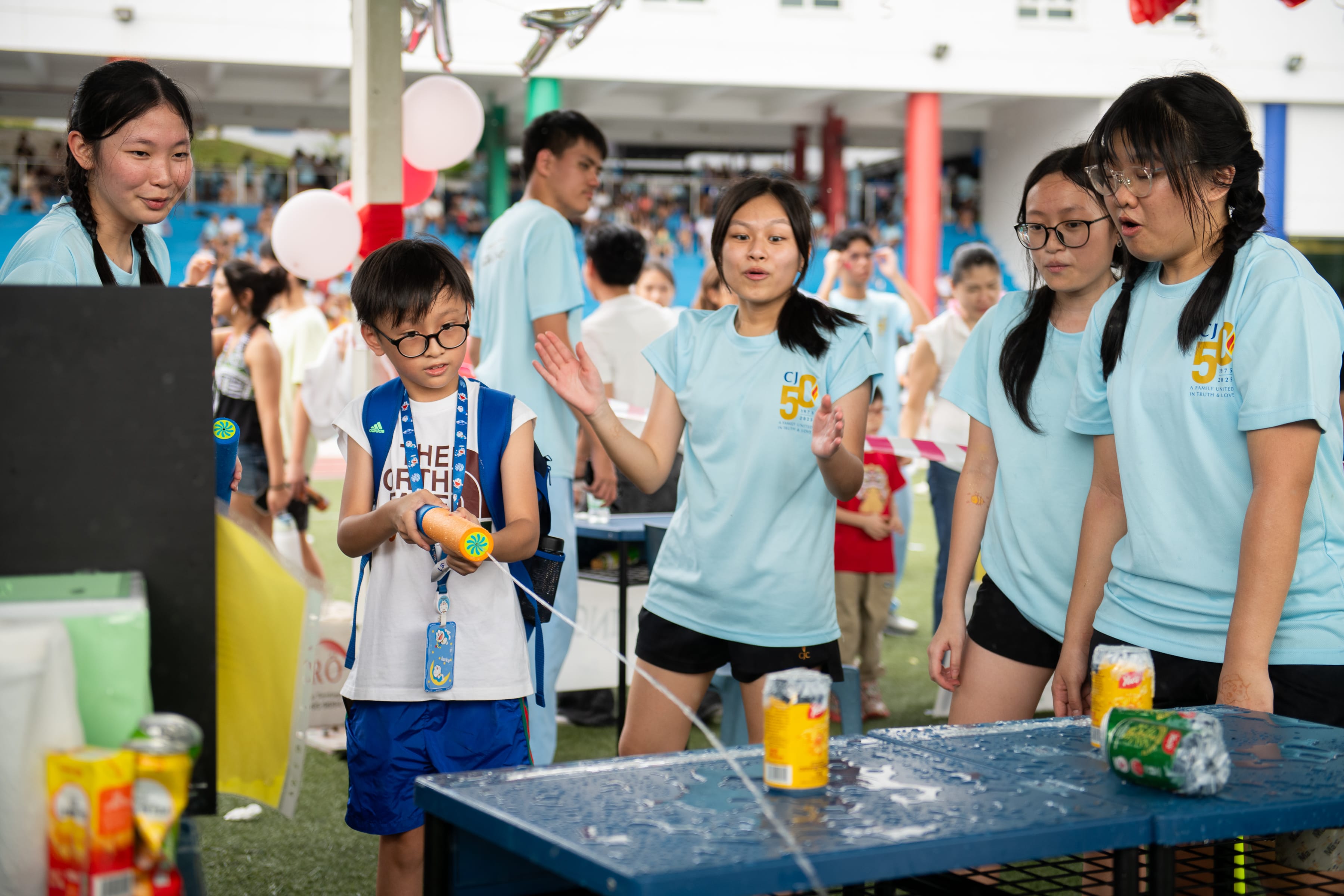 Boy with goggles aims a water blaster at cans on a table as four girls in blue shirts watch.