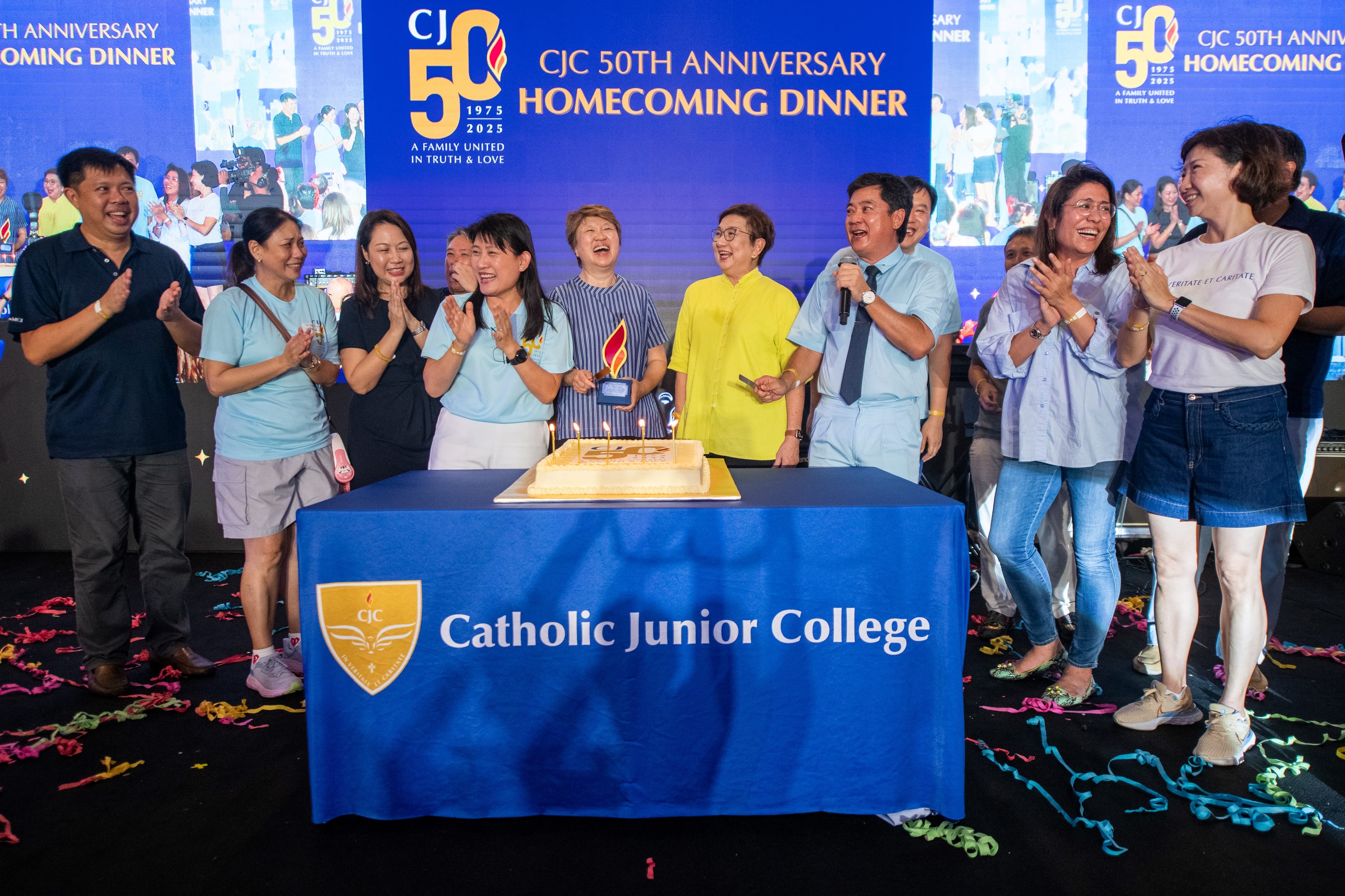 Group applauds around a lit cake at Catholic Junior College 50th anniversary homecoming dinner stage.