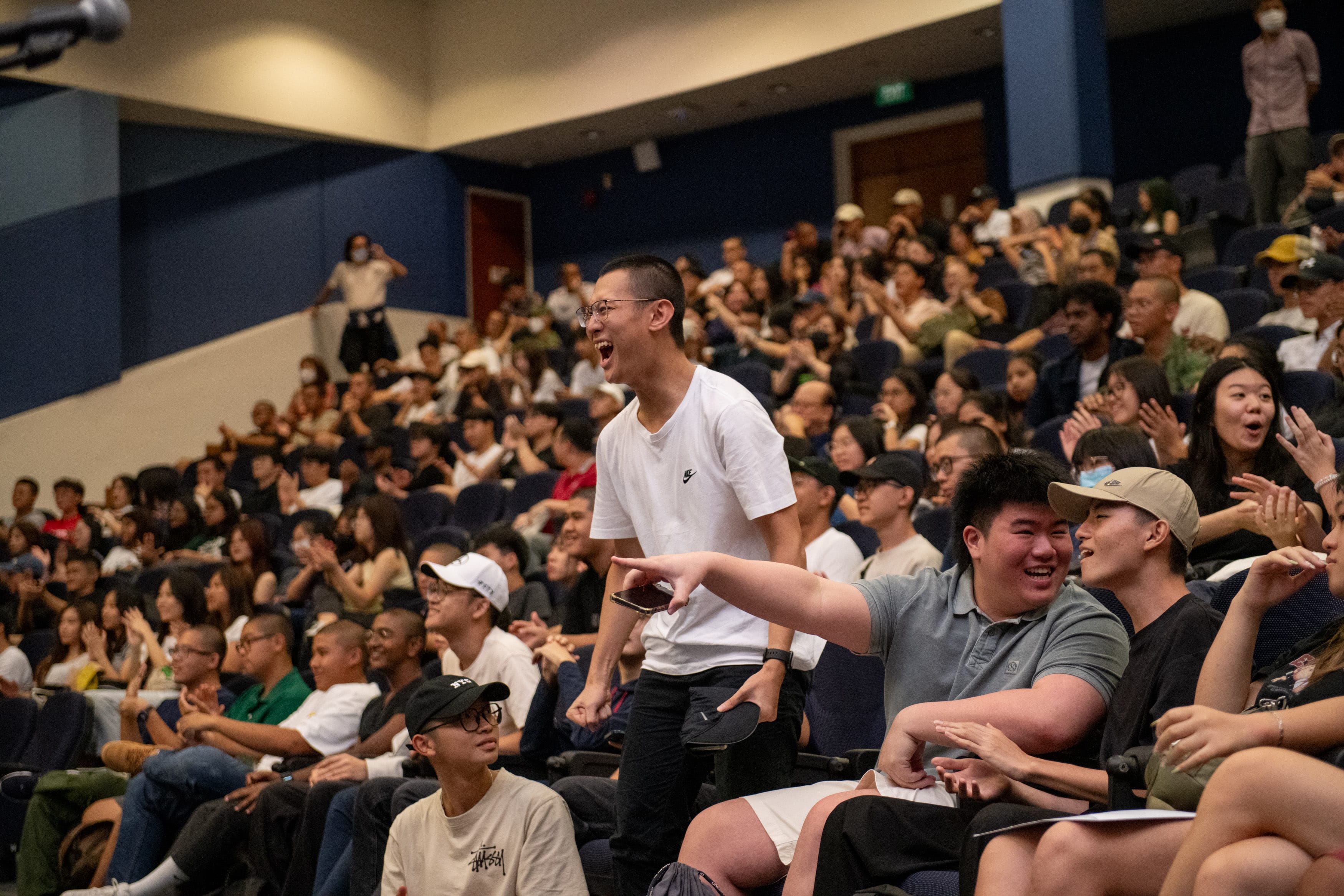A young man in a white Nike shirt stands and shouts excitedly in a packed auditorium full of cheering people.