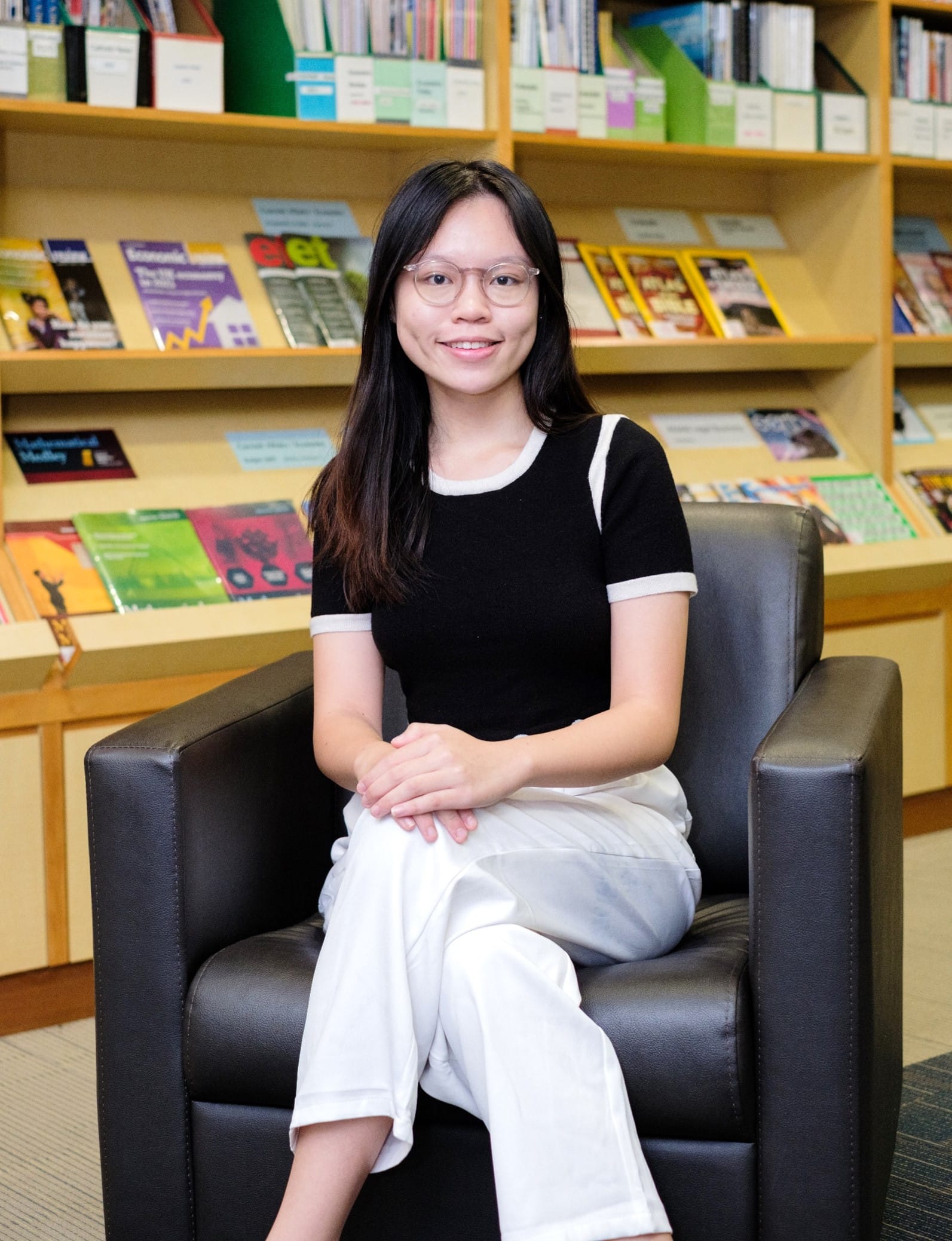 Young woman with glasses and long dark hair sits smiling in a black chair before wooden shelves of books.