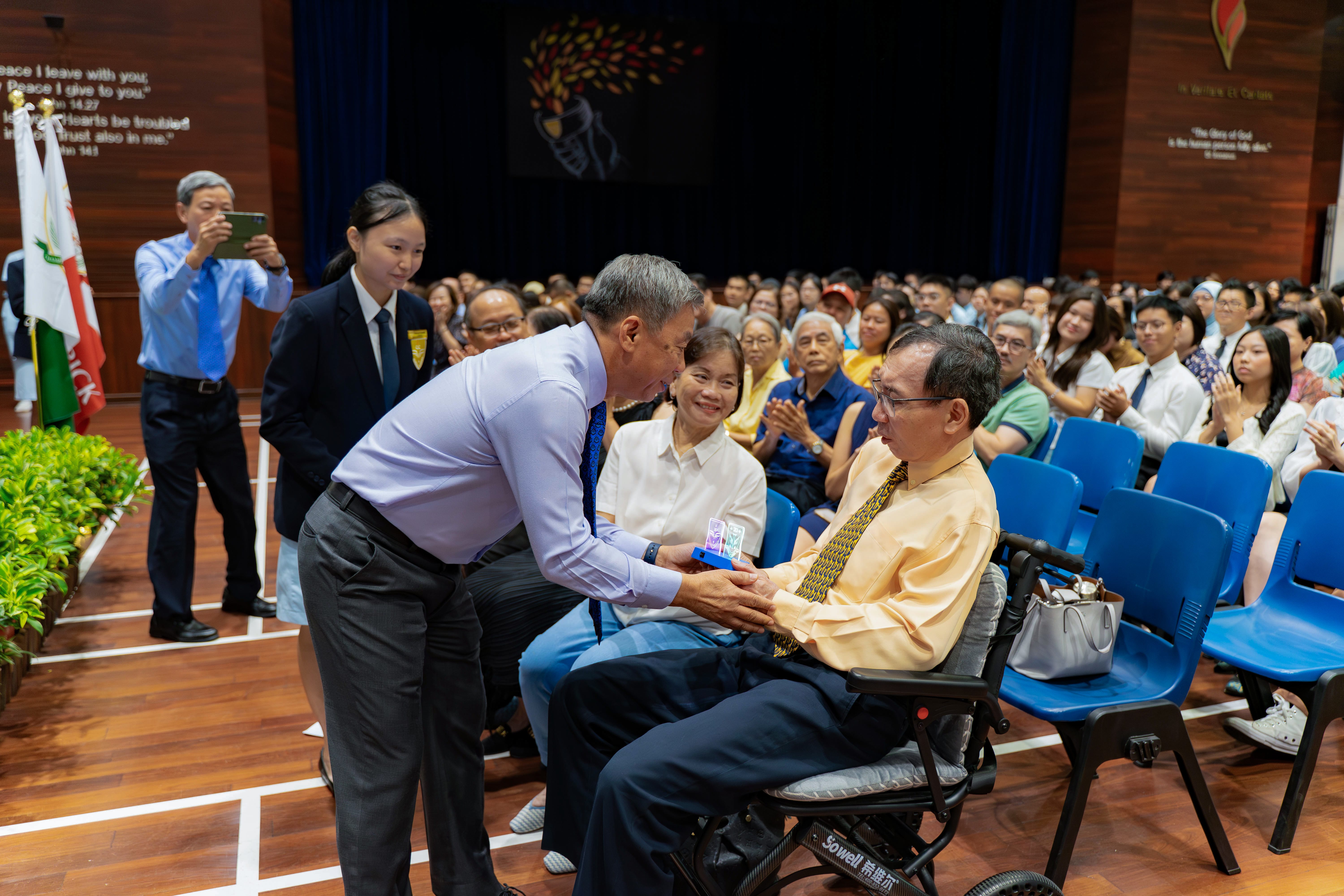 Man in purple shirt presents a small item to a seated man as an audience watches in an auditorium.