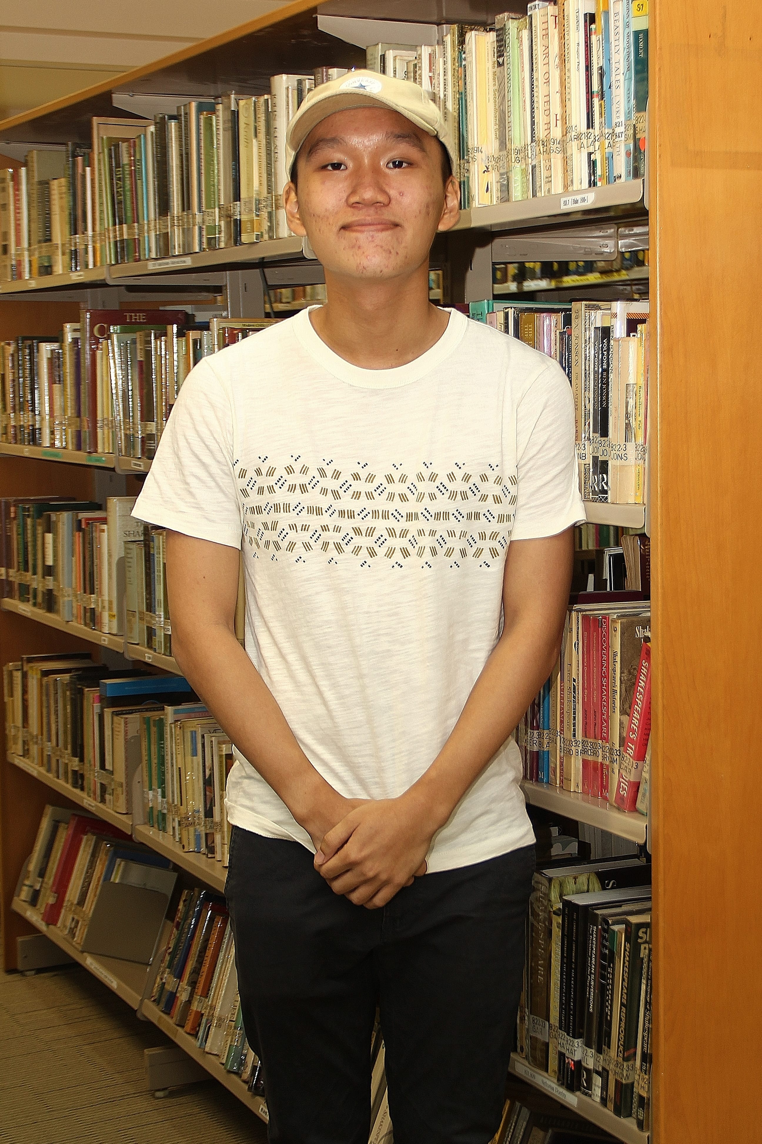 Person in a cap and white patterned T-shirt stands between library shelves filled with books.
