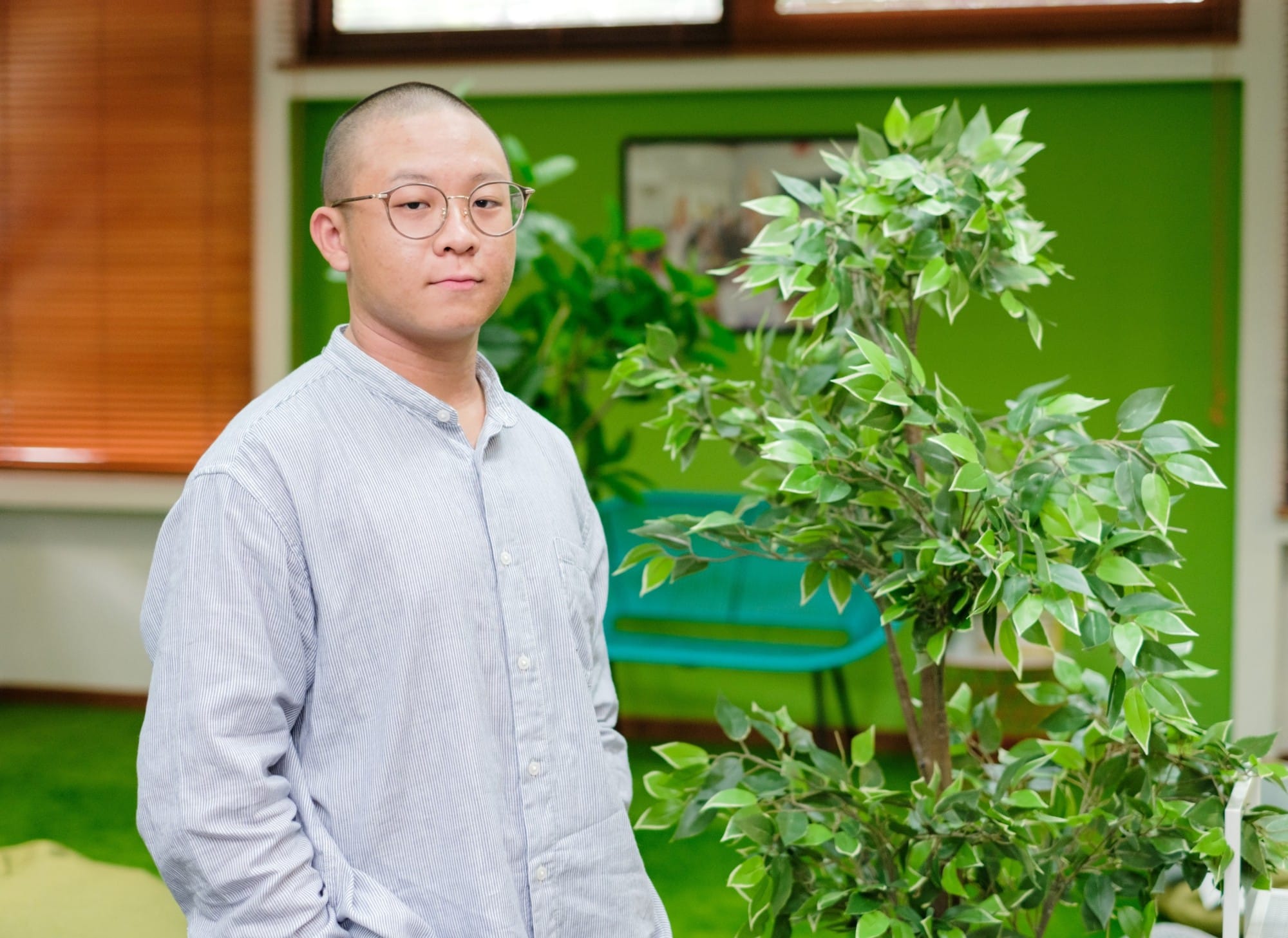 A young man with shaved head and round glasses wearing a light blue shirt stands beside a green leafy plant indoors.