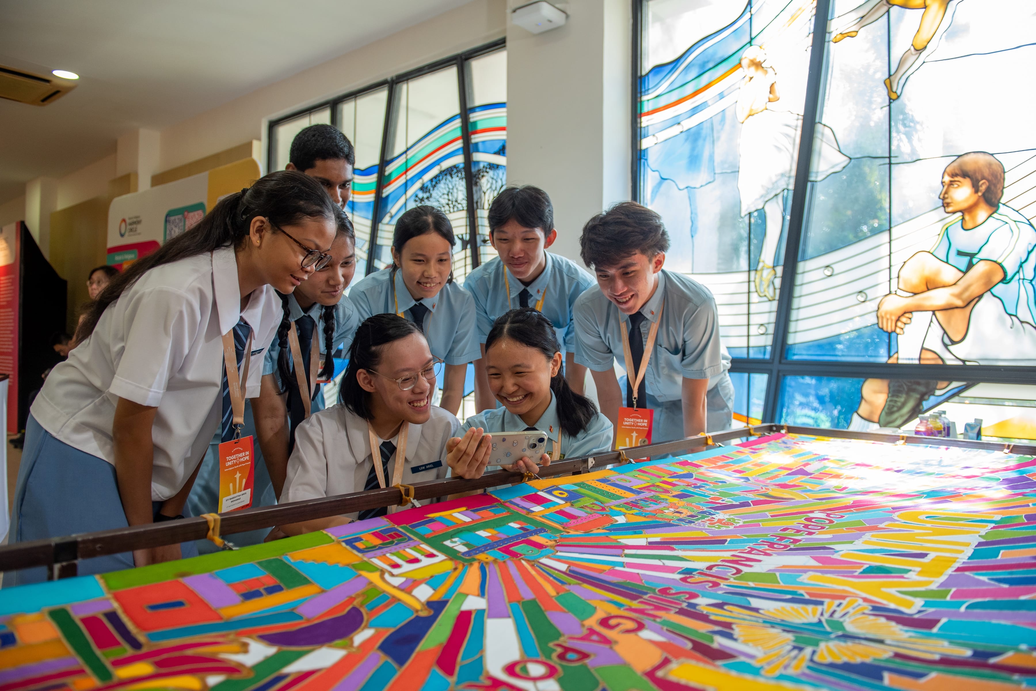 Group of students in uniforms gather smiling around a colorful table artwork, one holding a phone.